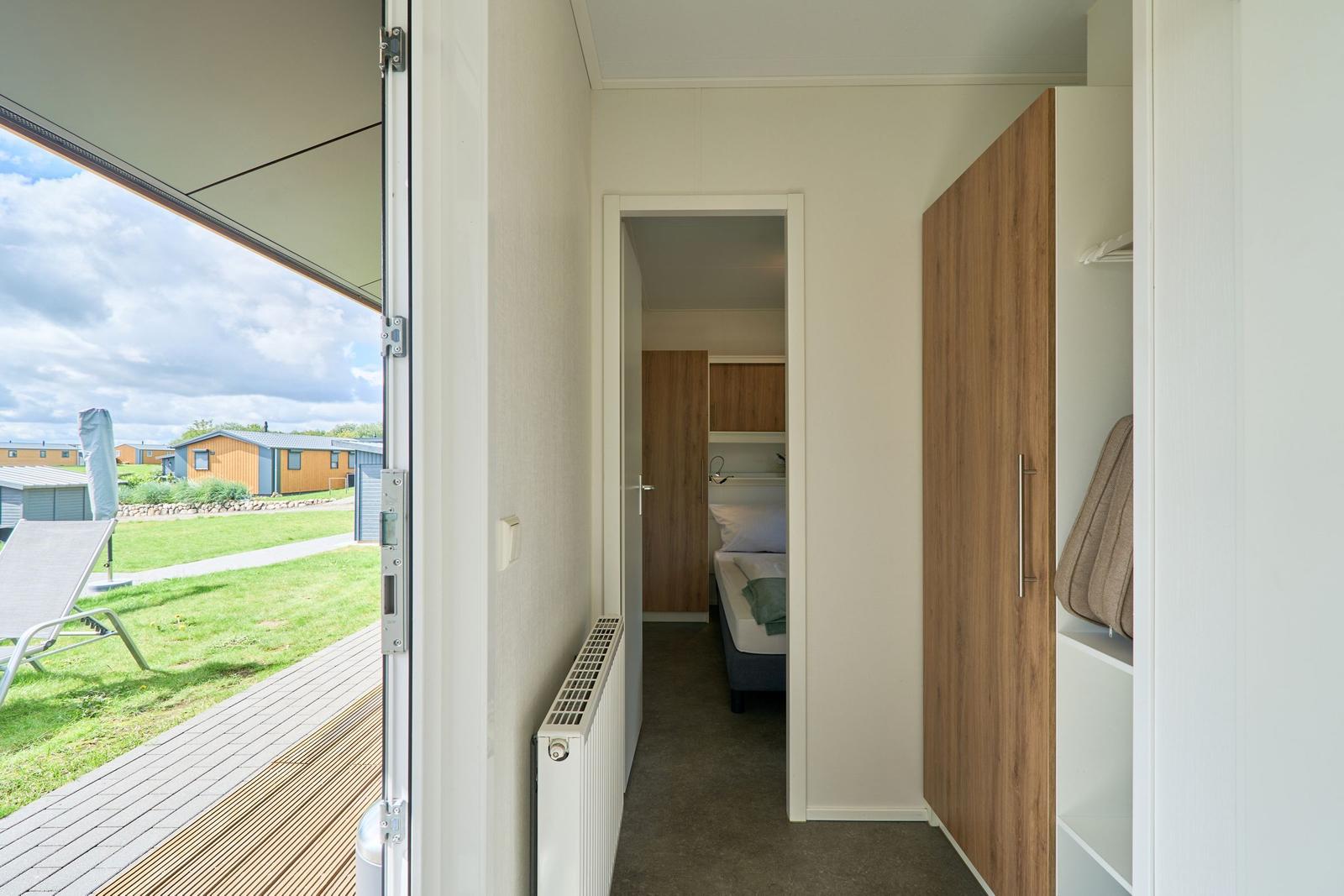 Schlafzimmer mit Bett und Kleiderschrank. Durchgang zur Terrasse mit Liegestuhl.