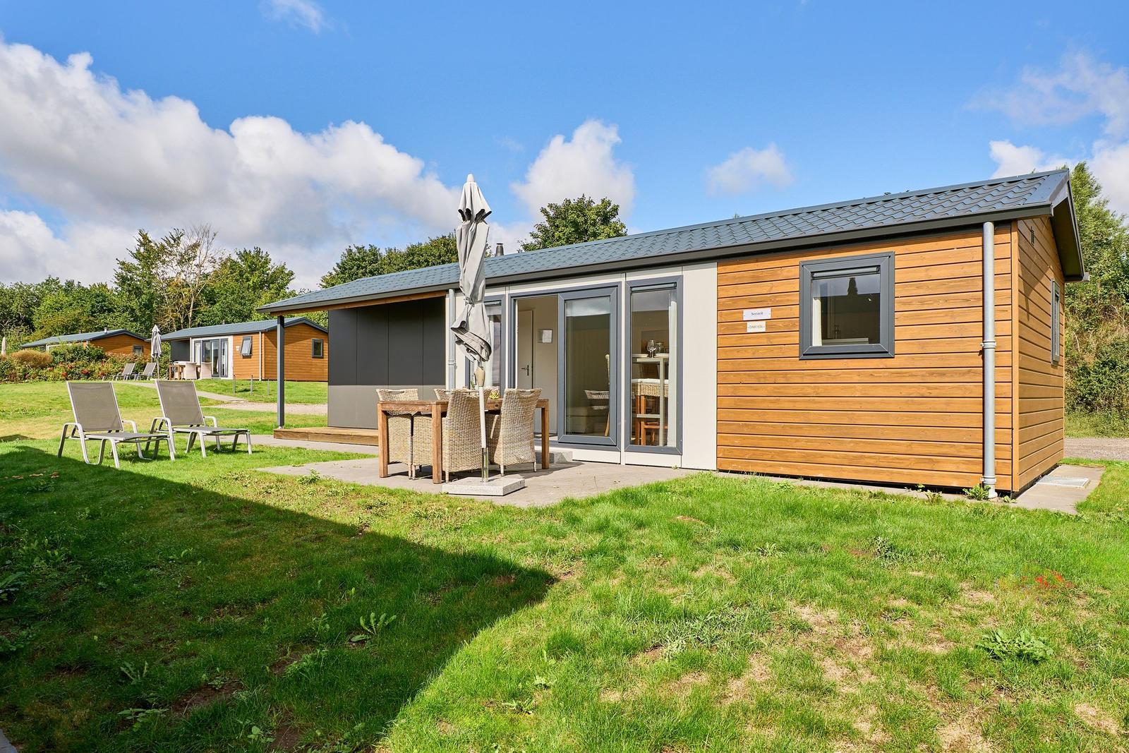 Holzhaus mit Terrasse, Garten und Außentisch. Hintergrund: weitere Häuser und Bäume.