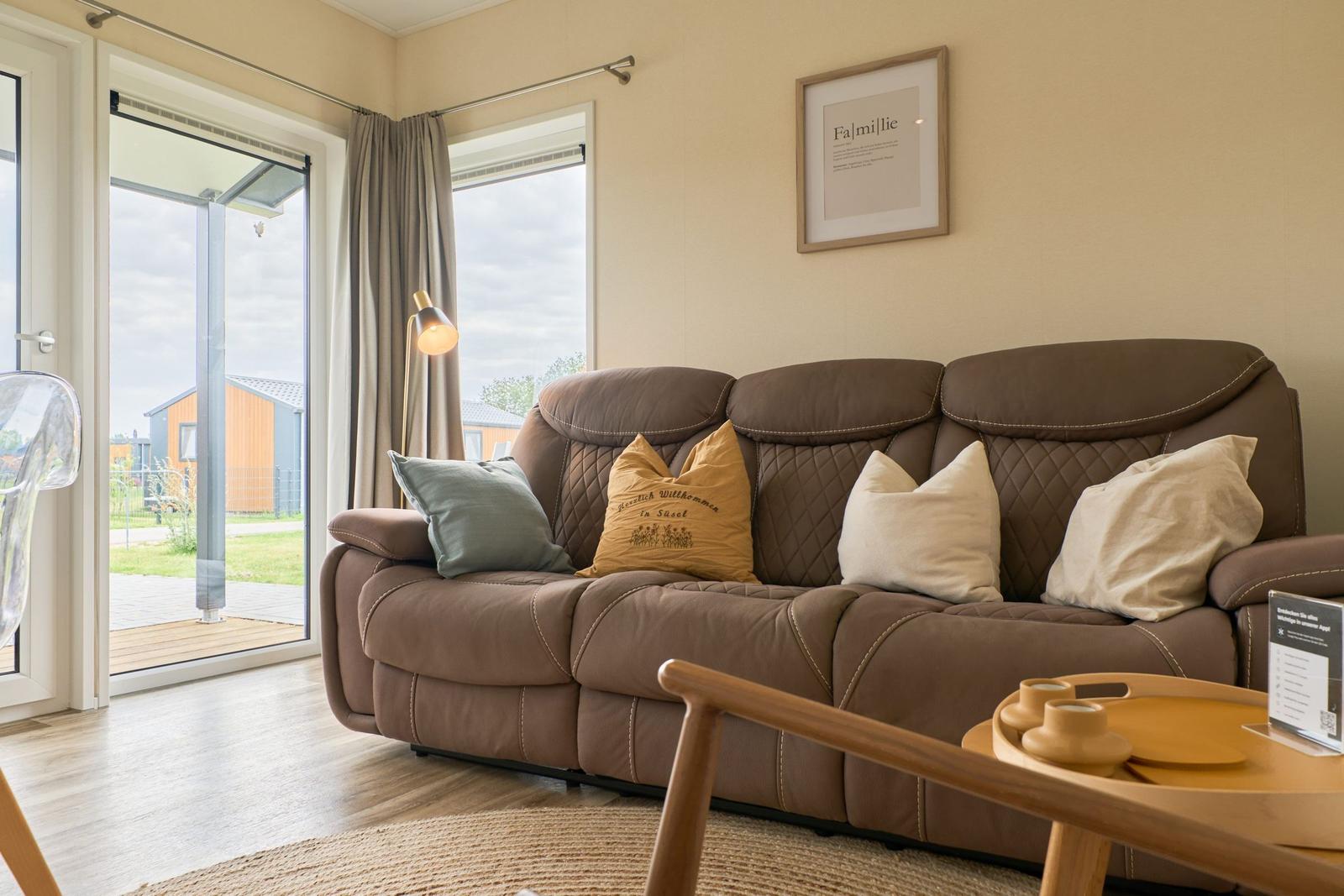 Ein braunes Sofa mit Kissen steht vor einem Fenster. Draußen ist eine Terrasse sichtbar.