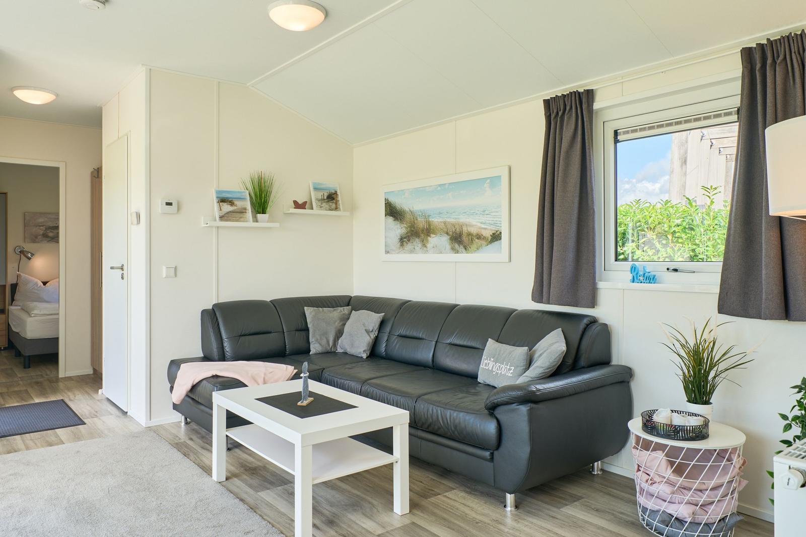 Wohnzimmer mit Sofa, Couchtisch und Fenster. Schlafzimmer im Hintergrund sichtbar.