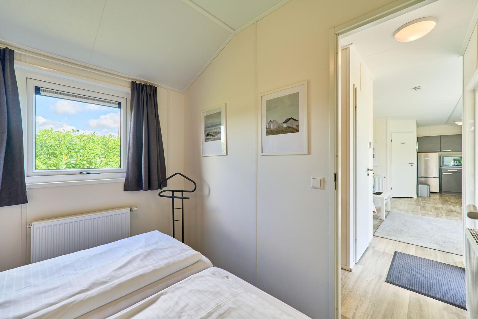 Schlafzimmer mit Fenster, Bett und Blick auf Grünflächen. Durchgang zur Küche.