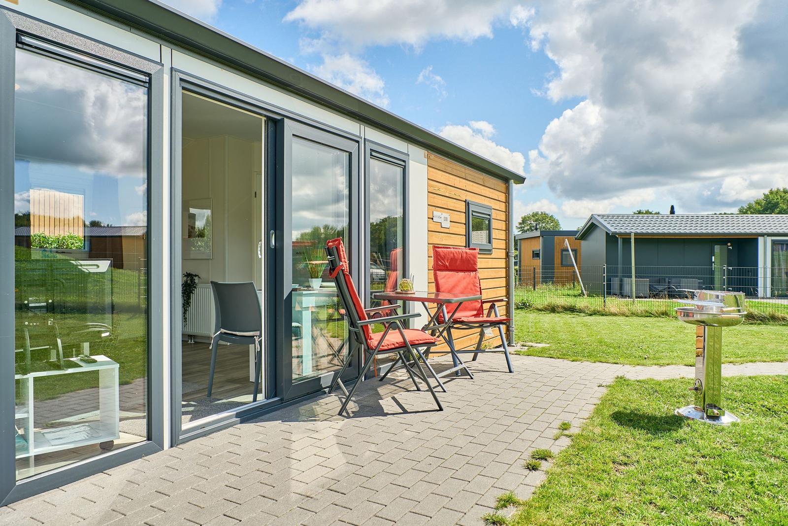 Terrasse mit Tisch, zwei roten Stühlen und Grill. Glasfront öffnet sich zum Innenraum.