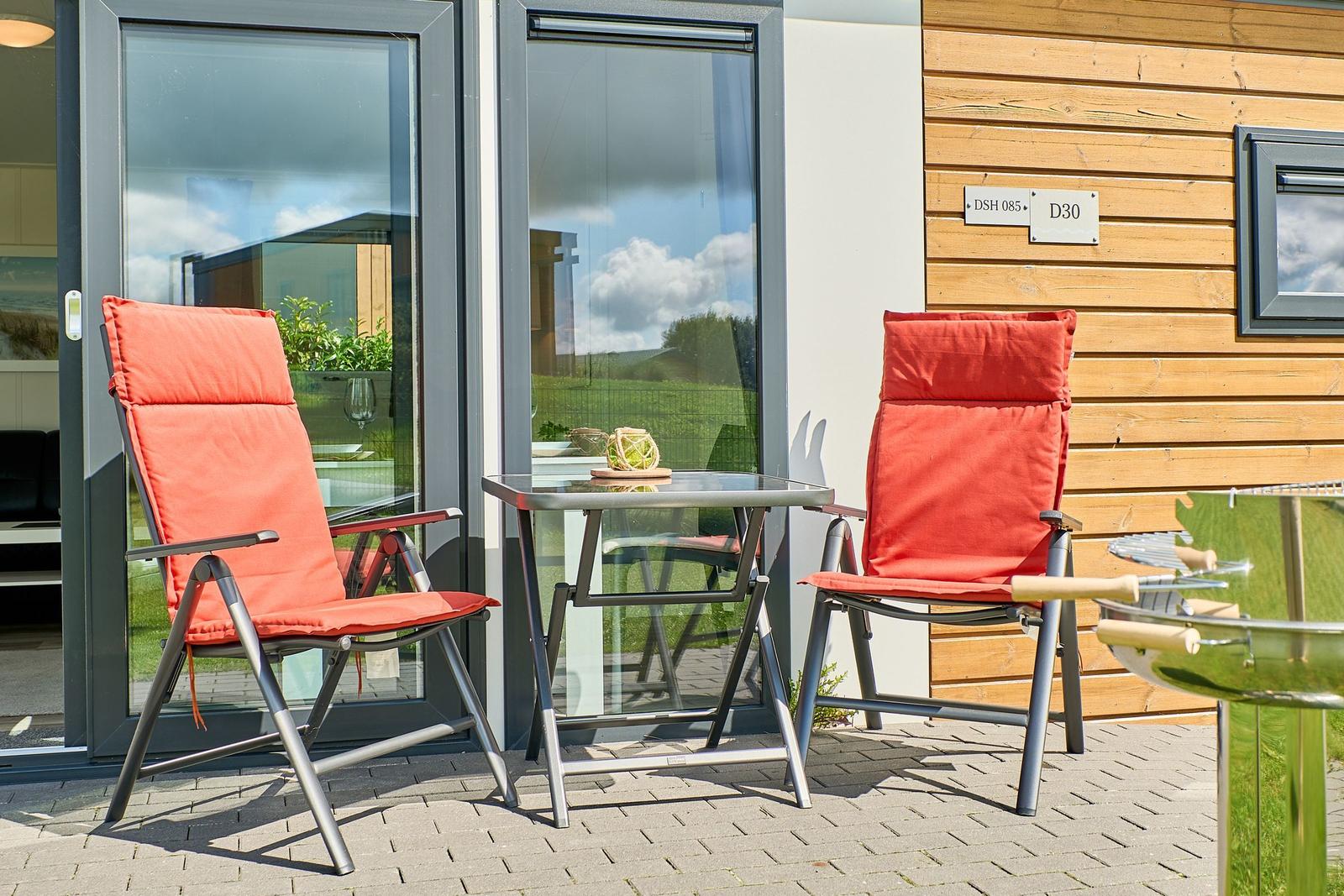 Terrasse mit zwei orangefarbenen Sesseln, Tisch und Grill. Hintergrund: Glasfront und Holzwand.
