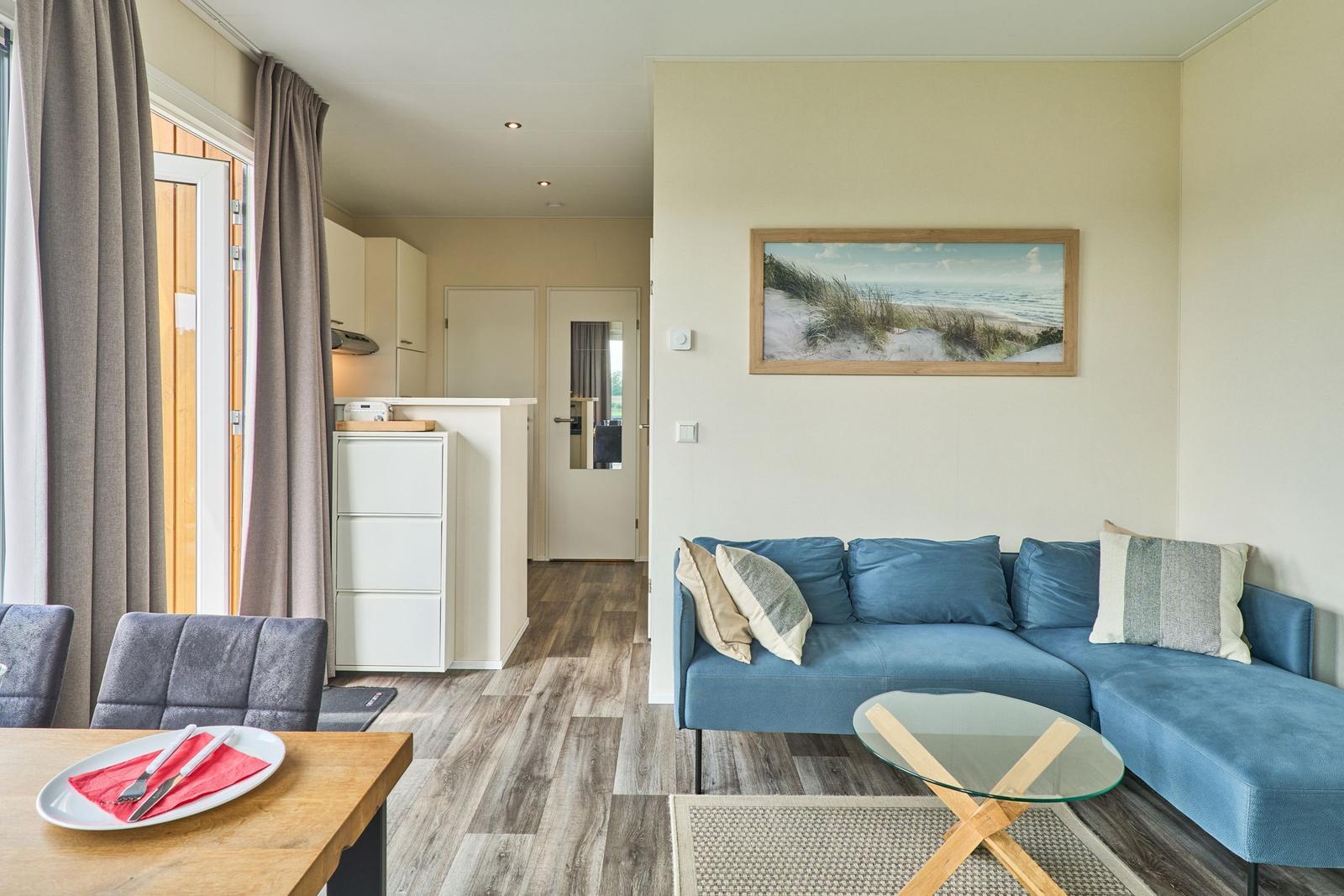 Wohn- und Essbereich mit blauem Sofa, Glas-Tisch und Küchenbereich im Hintergrund.