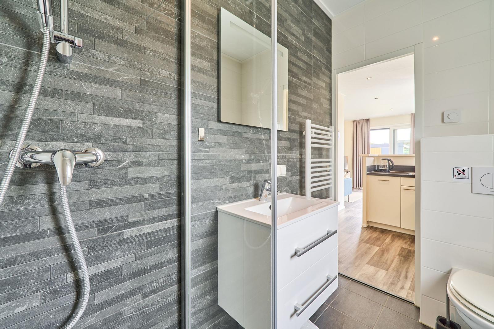 Badezimmer mit Dusche, Waschbecken und Toilette. Blick in die Küche.