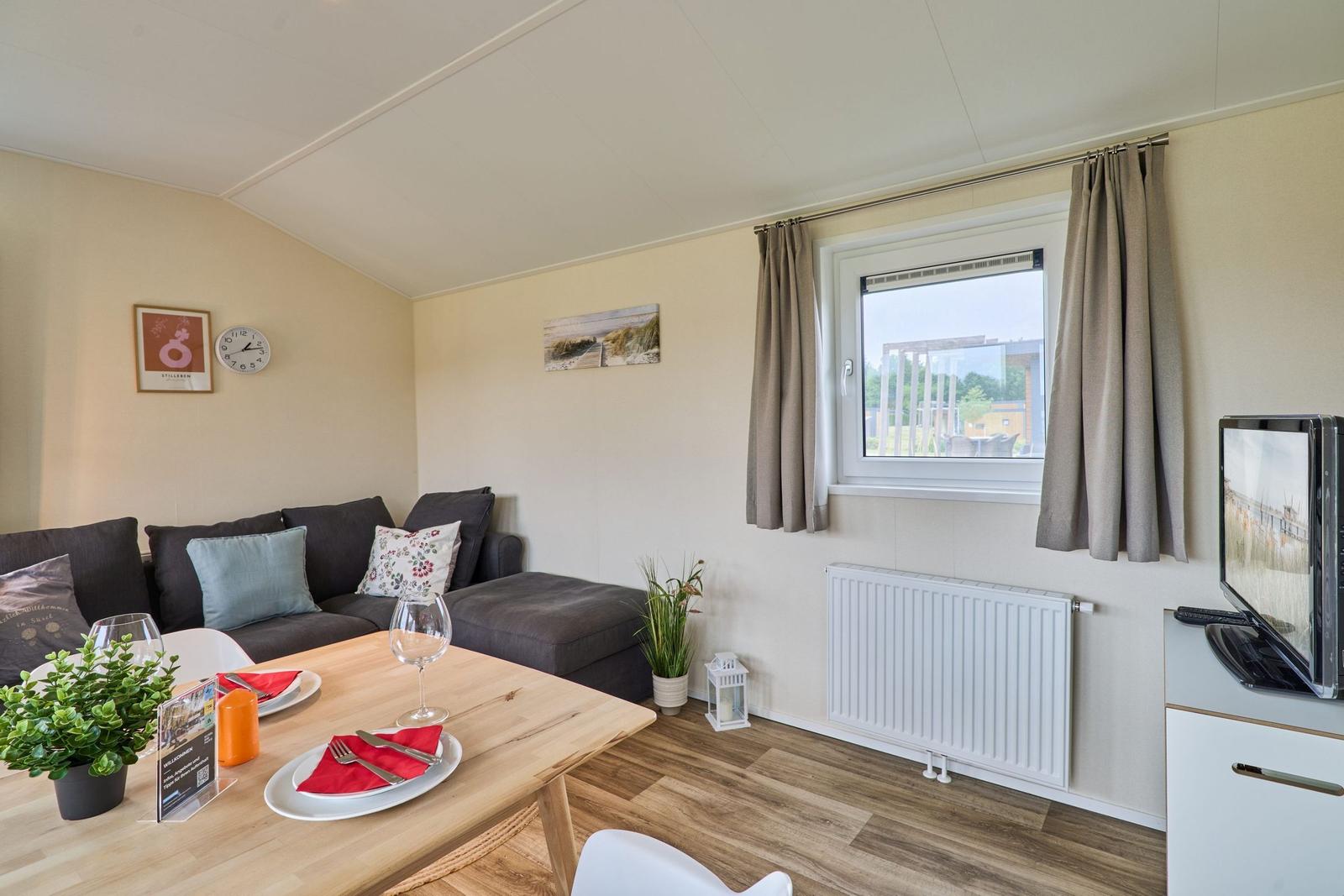 Wohnbereich mit Sofa, Esstisch und Fernseher. Fenster mit Vorhängen. Wandbilder und Uhr.