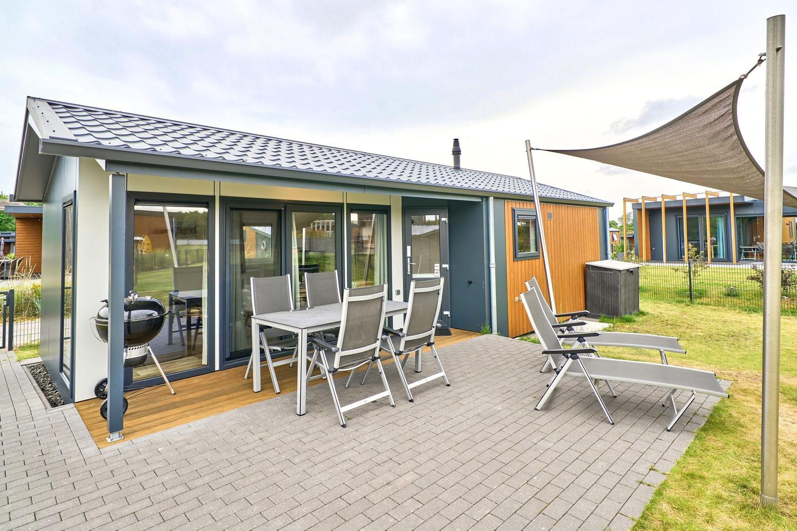 Terrasse mit Tisch, Stühlen, Liegestühlen und Grill. Sicht auf modernes Ferienhaus.