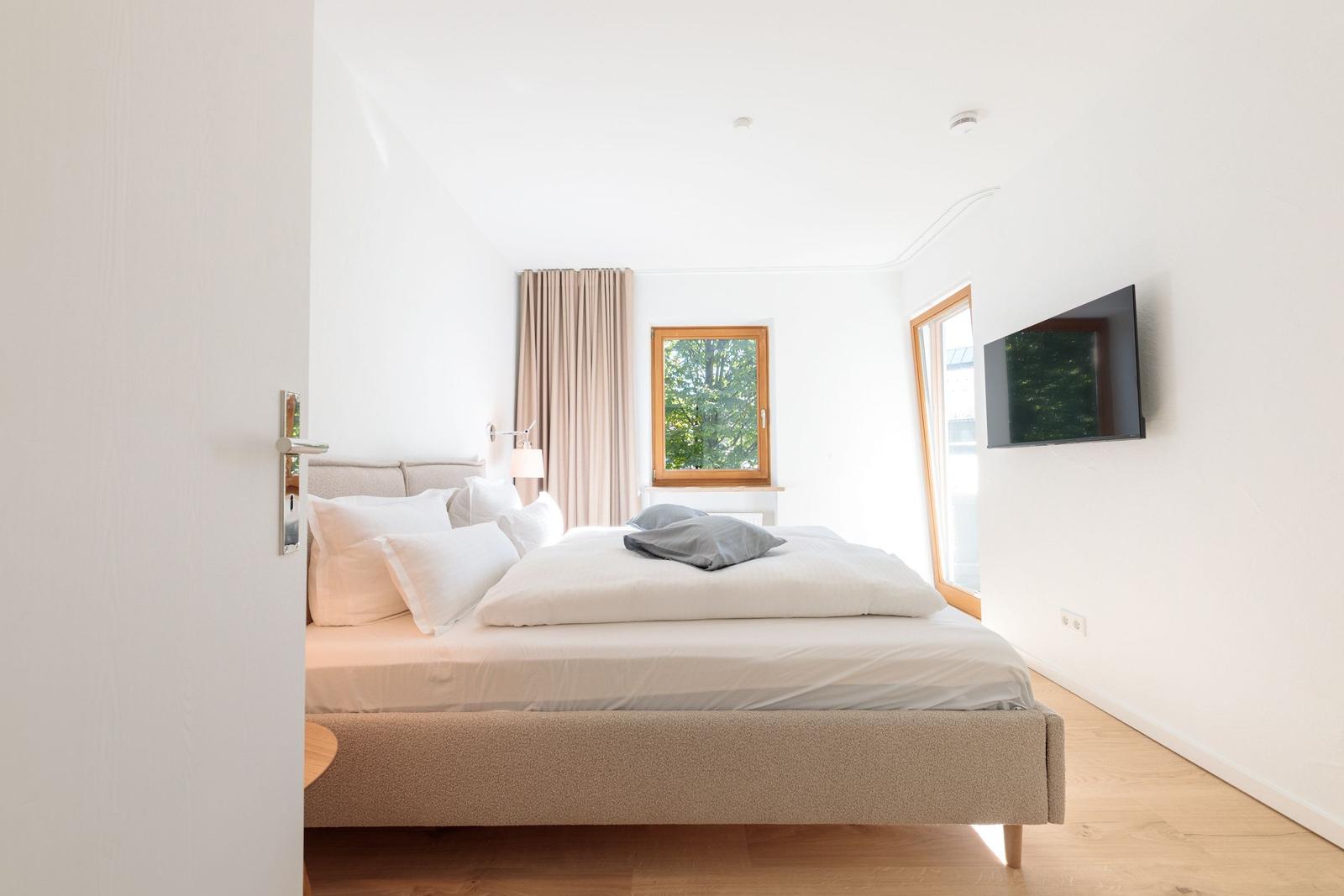 Schlafzimmer mit Bett, Fenstern und Wandfernseher.