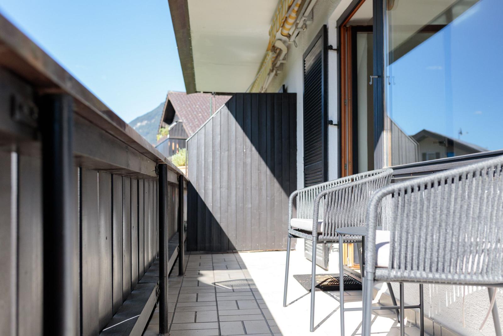 Terrasse mit zwei Stühlen und Tisch, Blick auf Gebäude und Berge.
