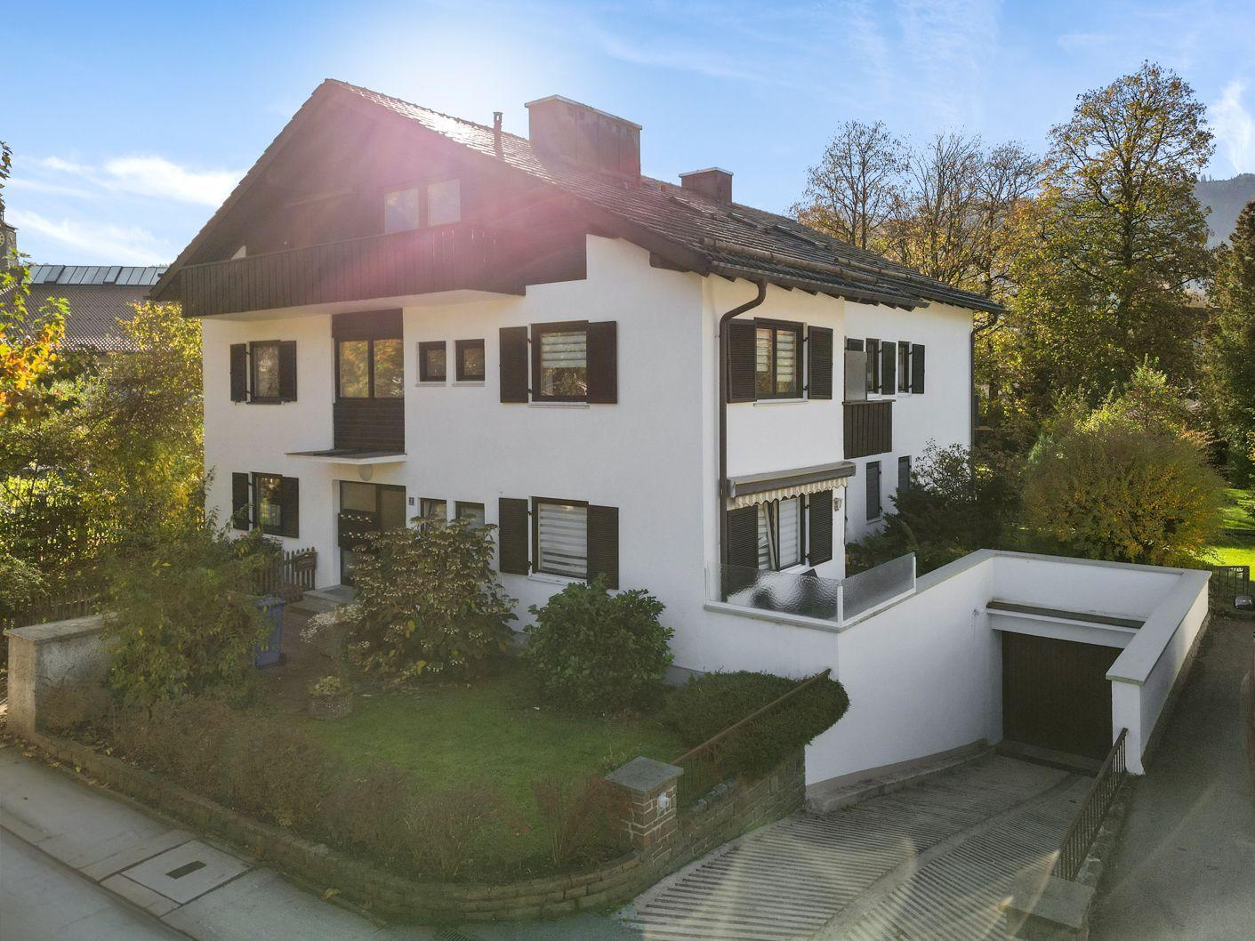 Ein zweistöckiges Haus mit weißen Wänden und dunklen Fensterläden. Umgeben von Grünflächen und Bäumen.