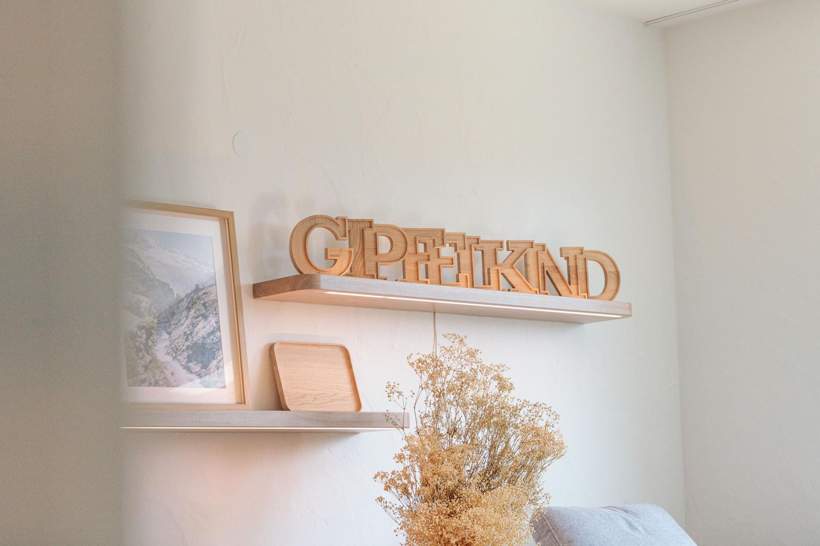 Wandregal mit Holz-Schriftzug und Bilderrahmen. Trockene Blumen im Vordergrund.