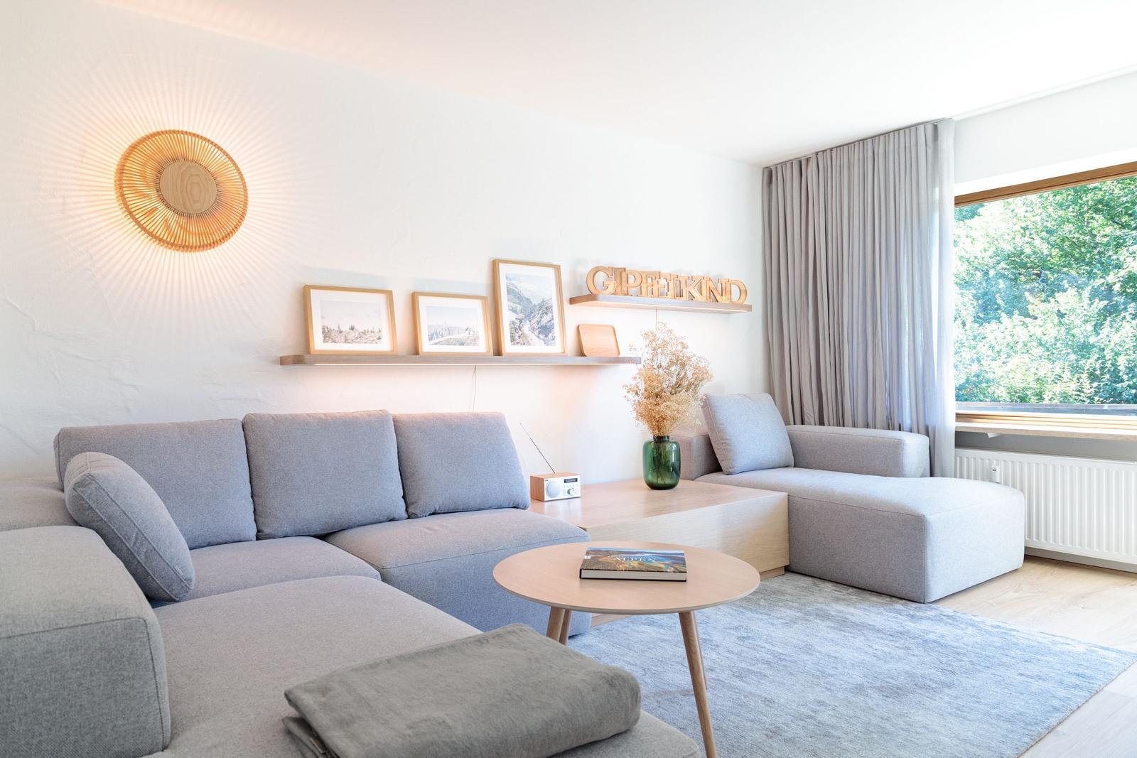 Ein Wohnzimmer mit grauem Sofa, Holztisch und großen Fenstern.