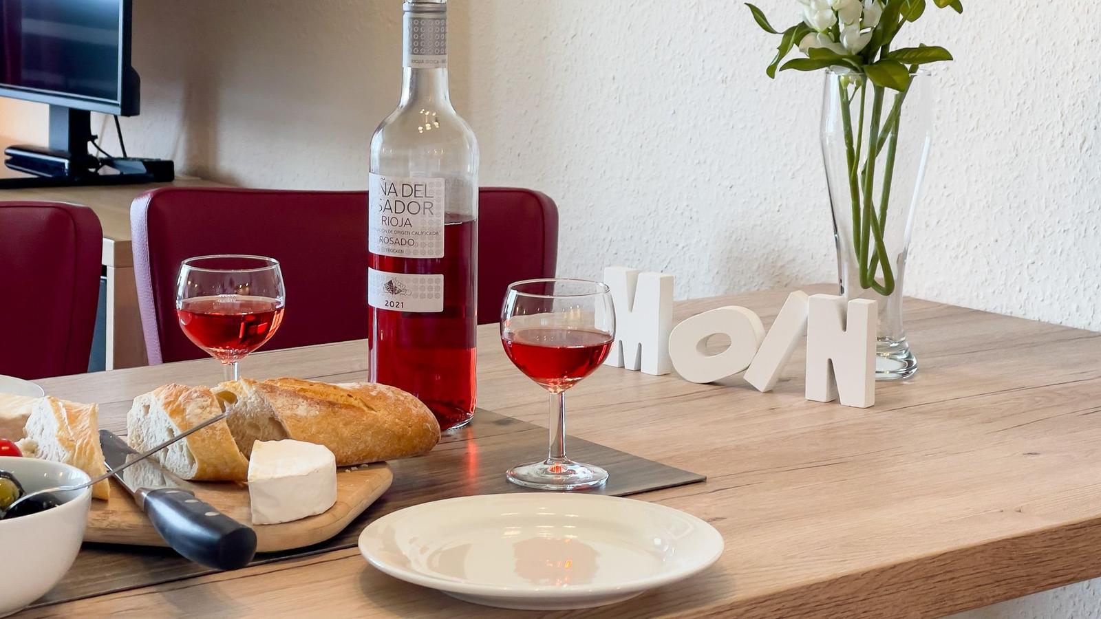 Tisch mit Wein, Brot und Käse, weiße Buchstaben und Blumen im Hintergrund.