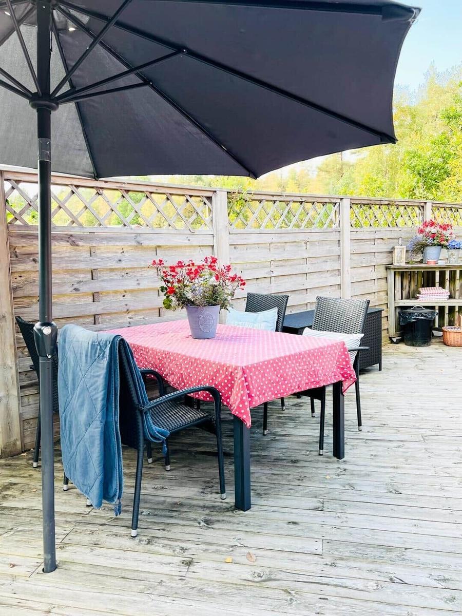 Terrasse mit Tisch, Stühlen und Sonnenschirm. Holzgeländer und Blumen sind sichtbar.