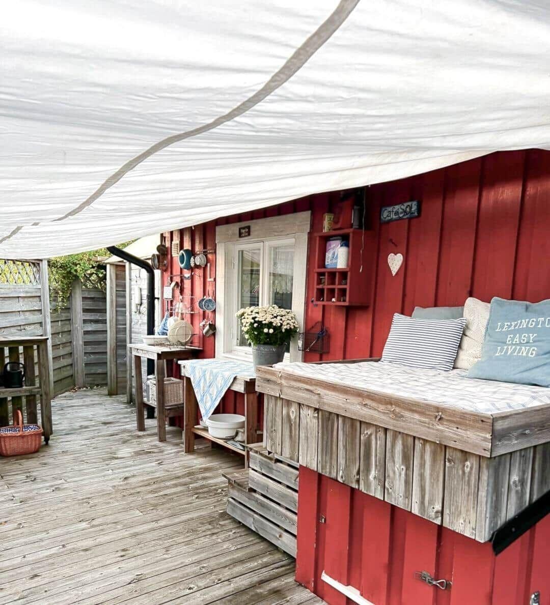 Roter Holzaussenbereich mit Sitzbank, Tisch und Blumen unter weißem Zelt.