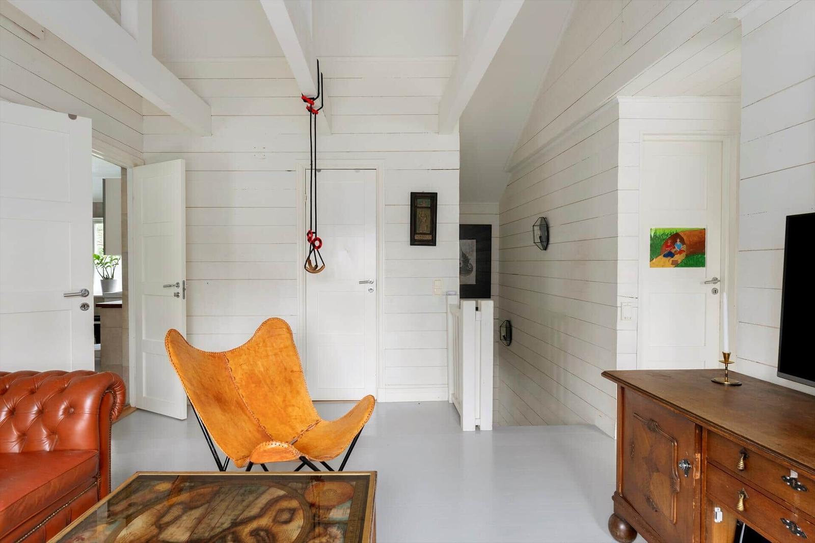 Wohnraum mit braunem Ledersofa, orangefarbenem Stuhl und Holzschrank. Weiße Wände und Deckenbalken.