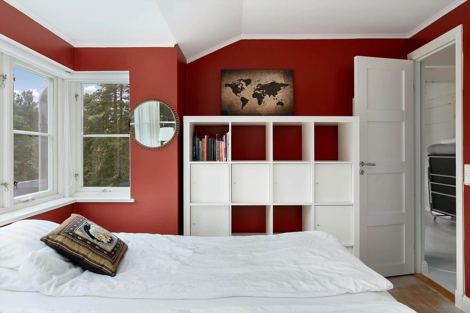 Schlafzimmer mit roten Wänden, weißem Bett und Bücherregal. Fenster mit Blick auf Wald.