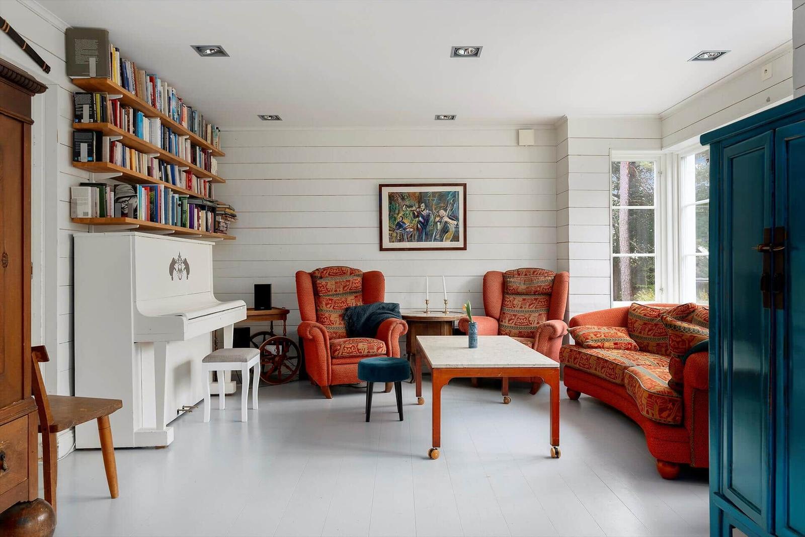 Wohnzimmer mit Klavier, Bücherregal und orangefarbenen Sitzmöbeln.