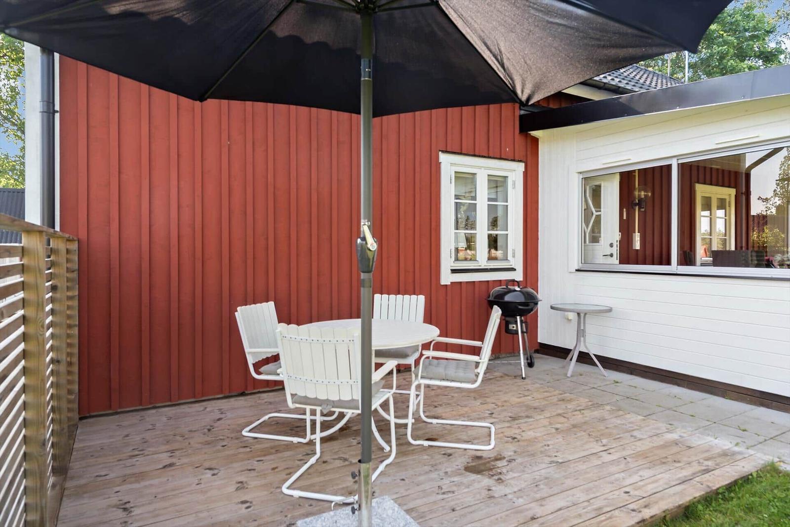 Terrasse mit Tisch, Stühlen, Sonnenschutz und Grill vor rotem und weißem Haus.