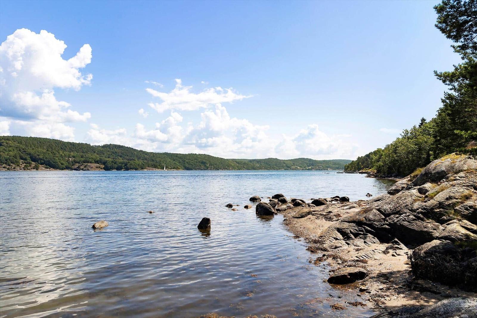 Ruhiger See mit Felsenstrand und bewaldeten Hügeln unter blauem Himmel.