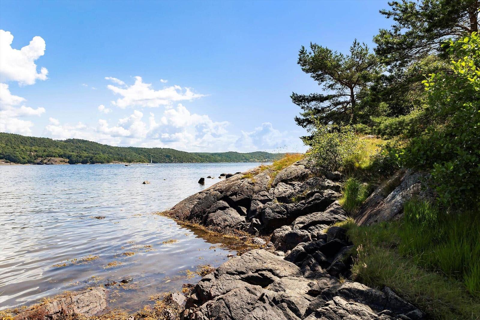 Steiniger Uferbereich mit Blick auf ruhiges Wasser und bewaldete Inseln unter blauem Himmel.