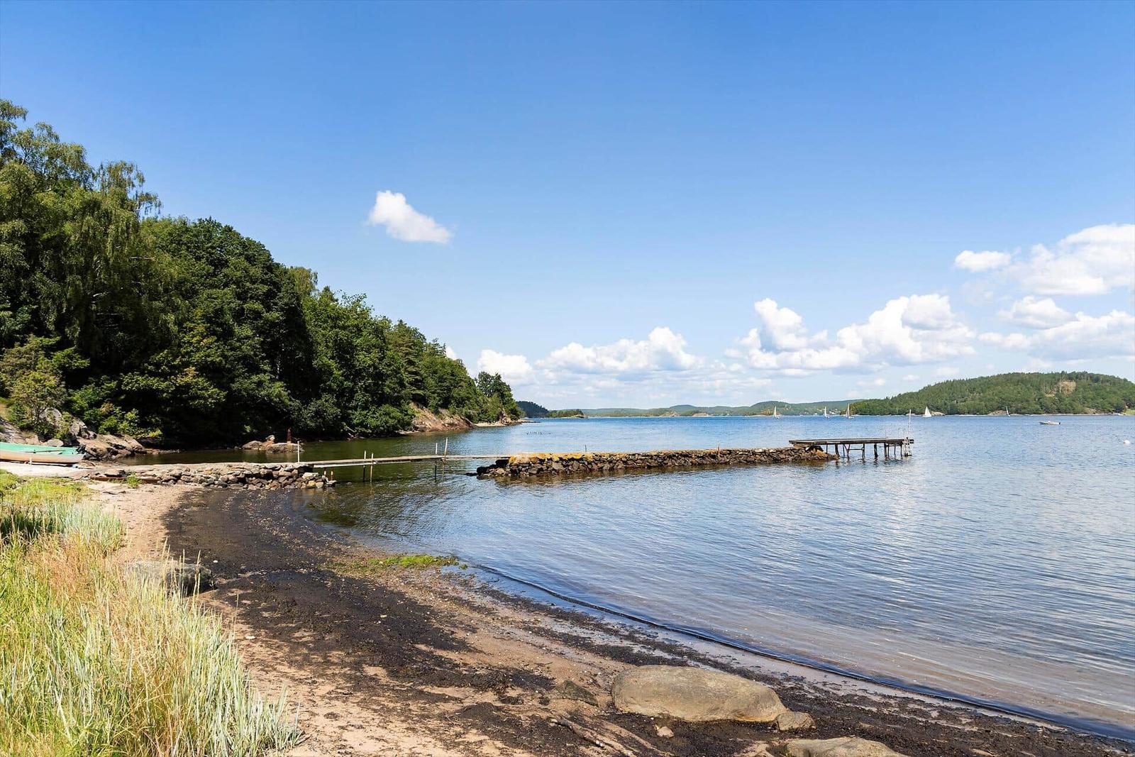 Strand mit Steg und Booten an ruhigem See unter blauem Himmel.