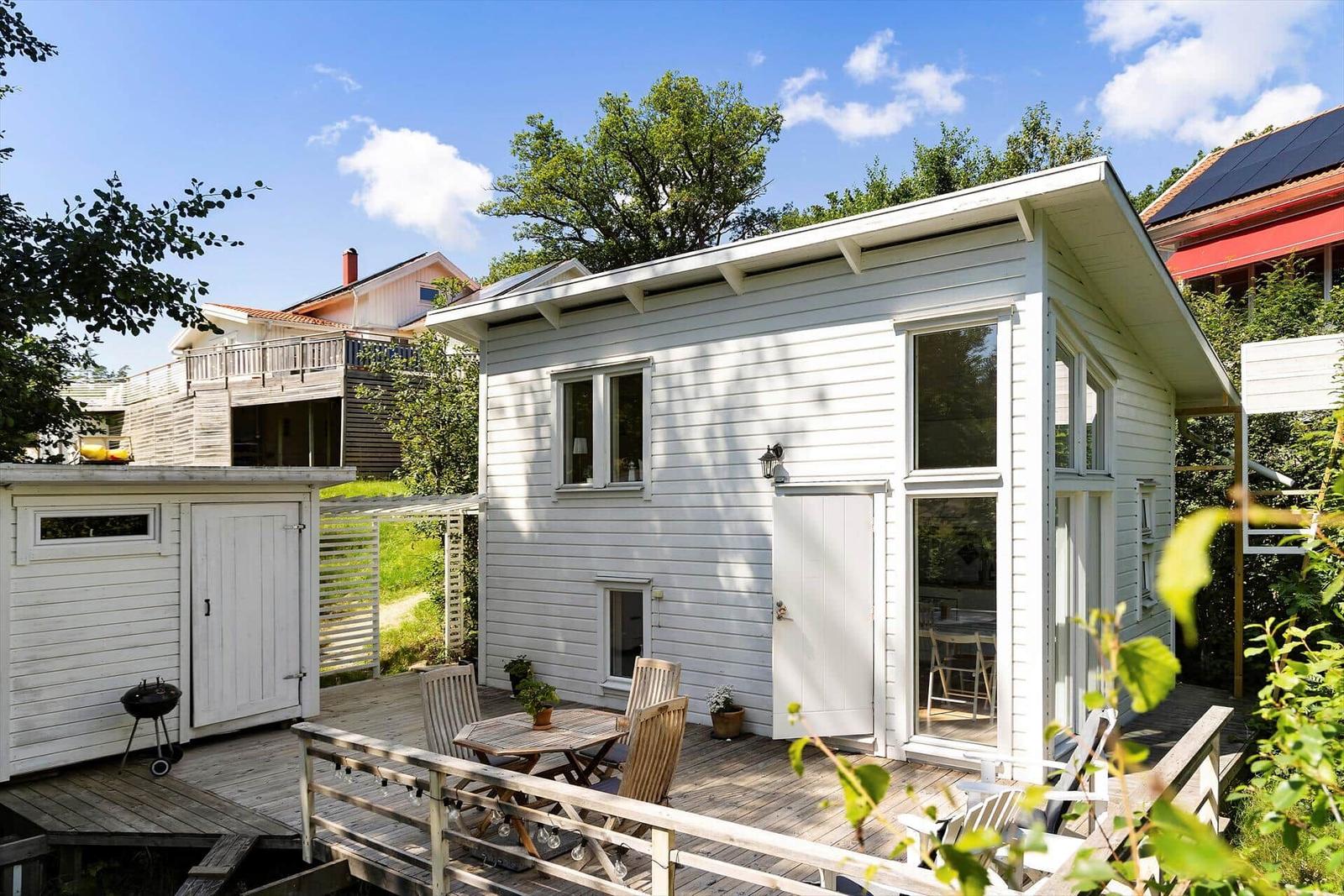 Weiße Holzhütte mit Terrasse, Tisch und Stühlen. Grillhaus und Garten im Hintergrund.