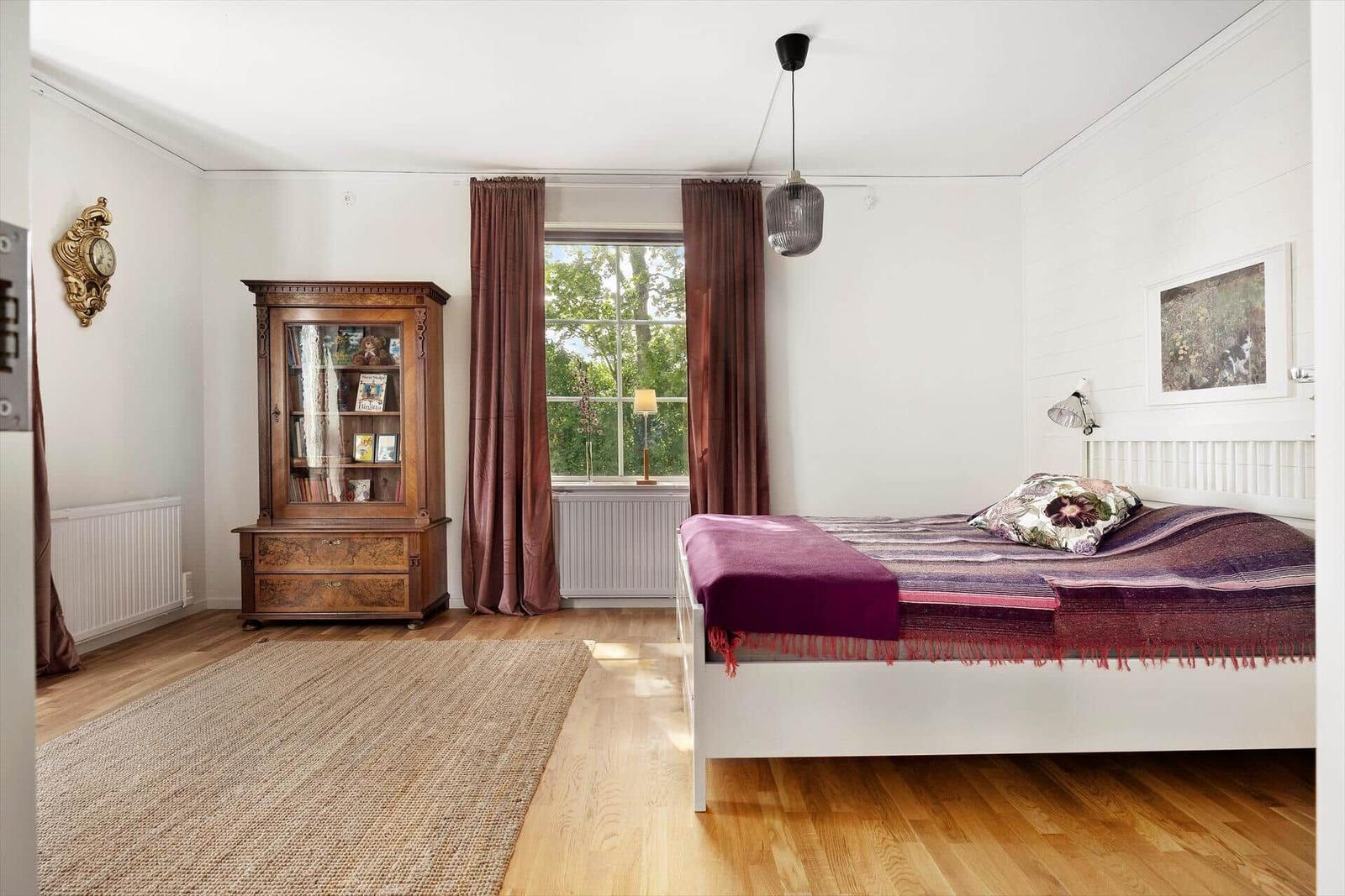 Schlafzimmer mit weißem Bett, Holzboden und Holzschrank. Fenster mit braunen Vorhängen.