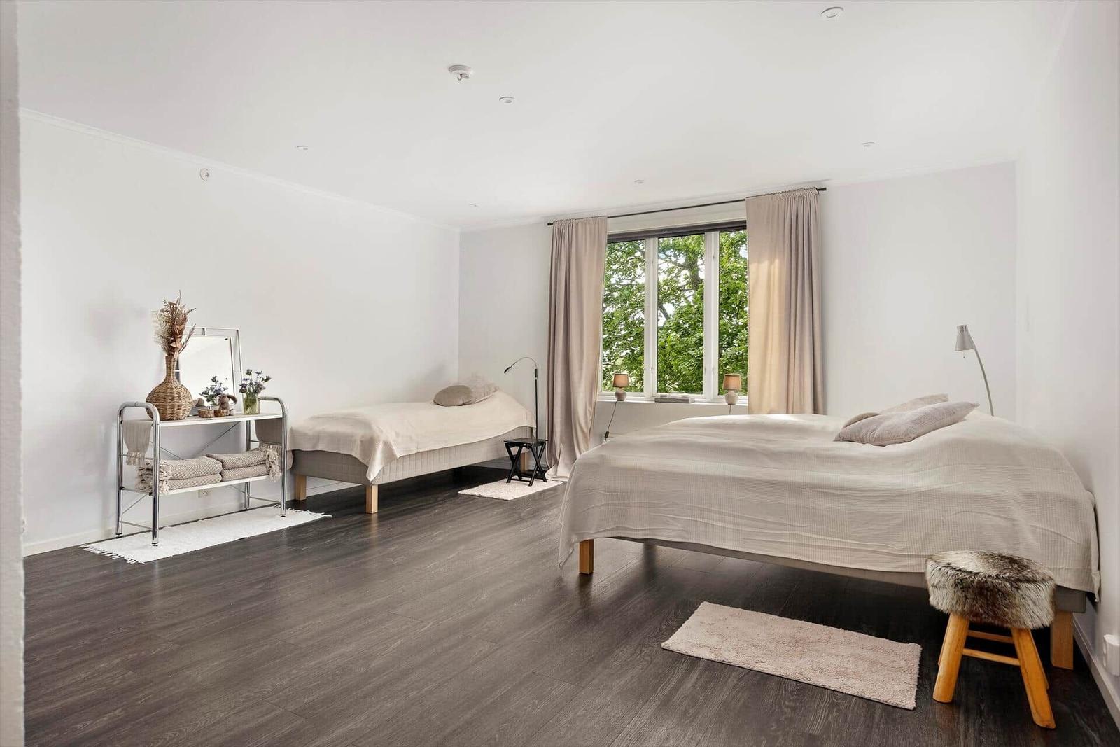 Zwei Betten, Fenster mit Blick auf Bäume, dunkler Holzboden und einfache Möbel.