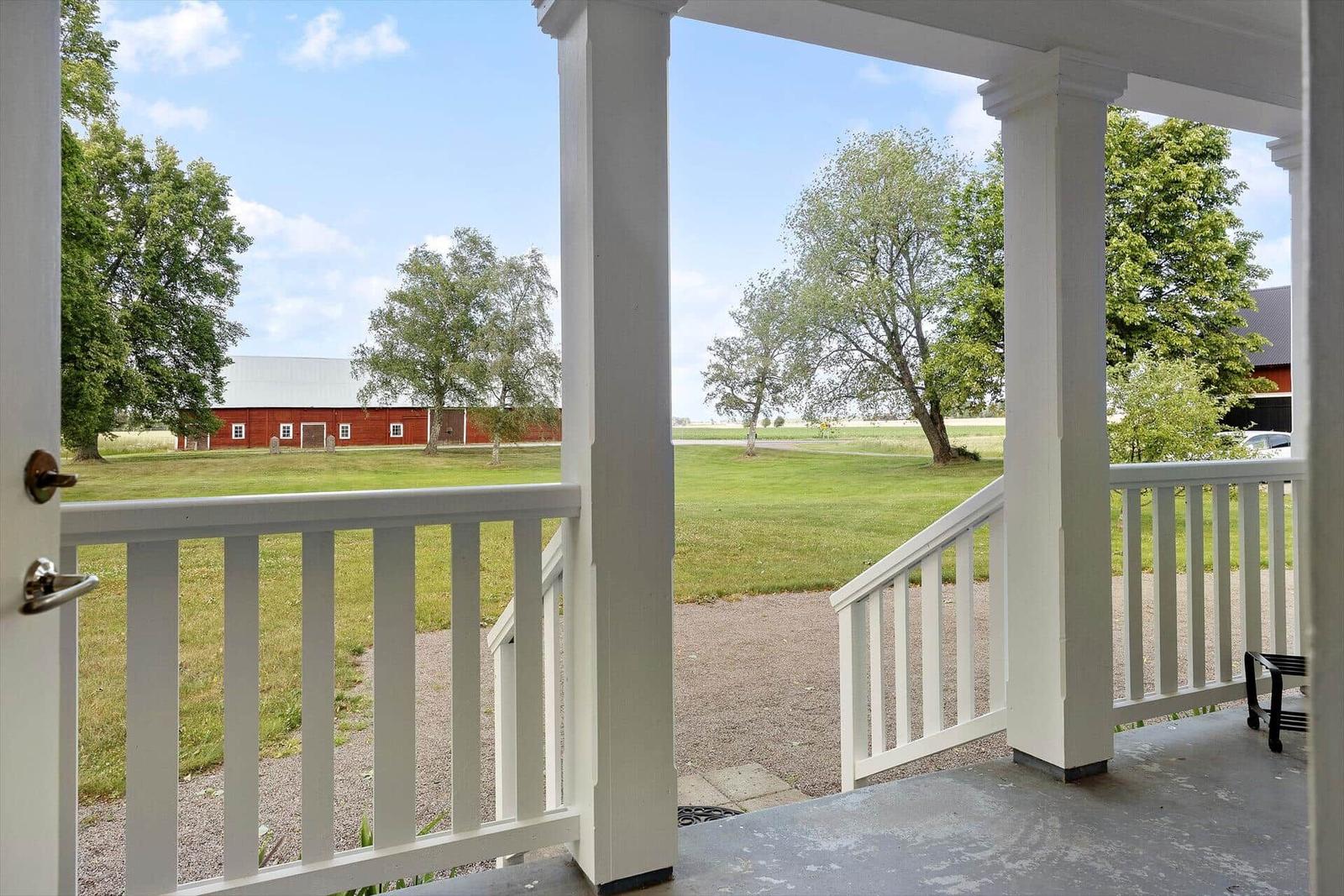 Vor dem Haus: weißer Veranda mit Sicht auf rote Scheune und grünes Feld.