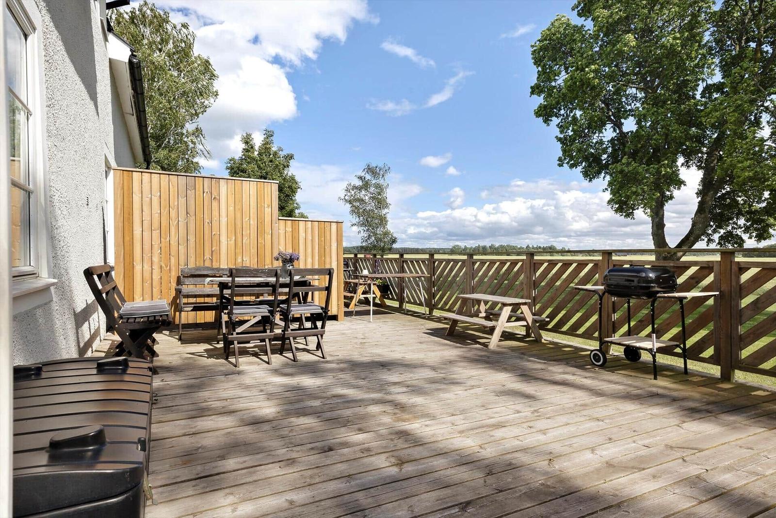 Holzterrasse mit Tisch, Stühlen, Picknicktisch und Grill. Blick auf grüne Landschaft.