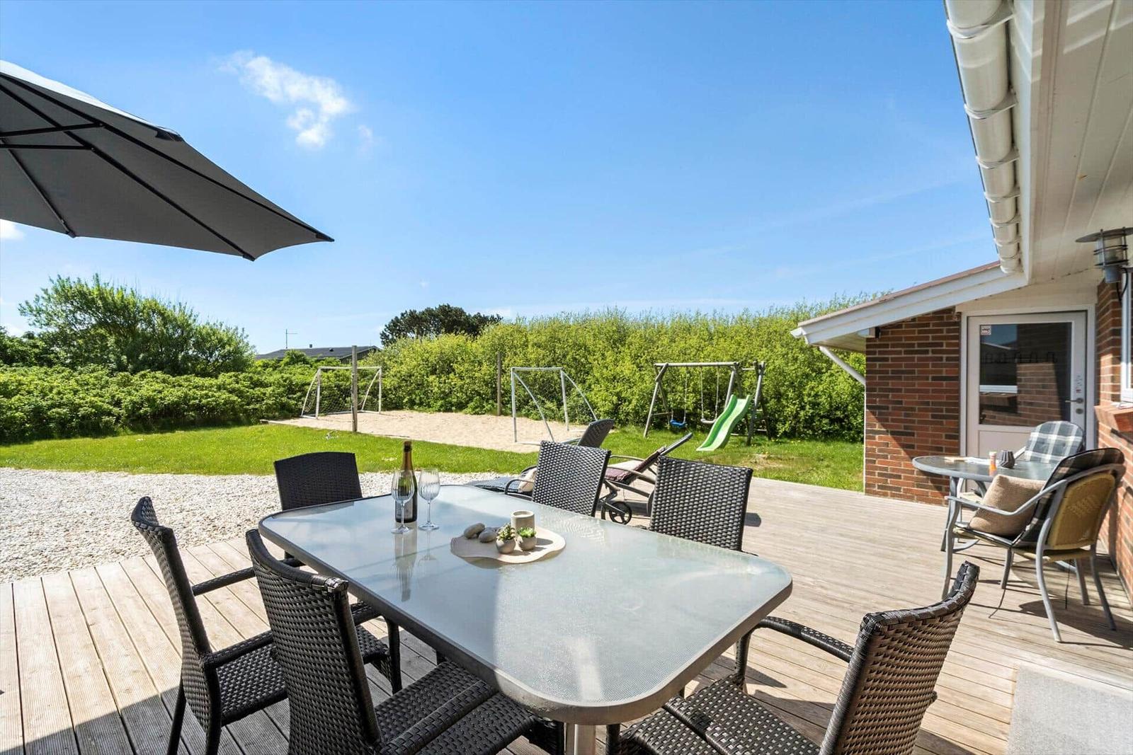 Deck with table, chairs, and umbrella. Playground and green area in background.