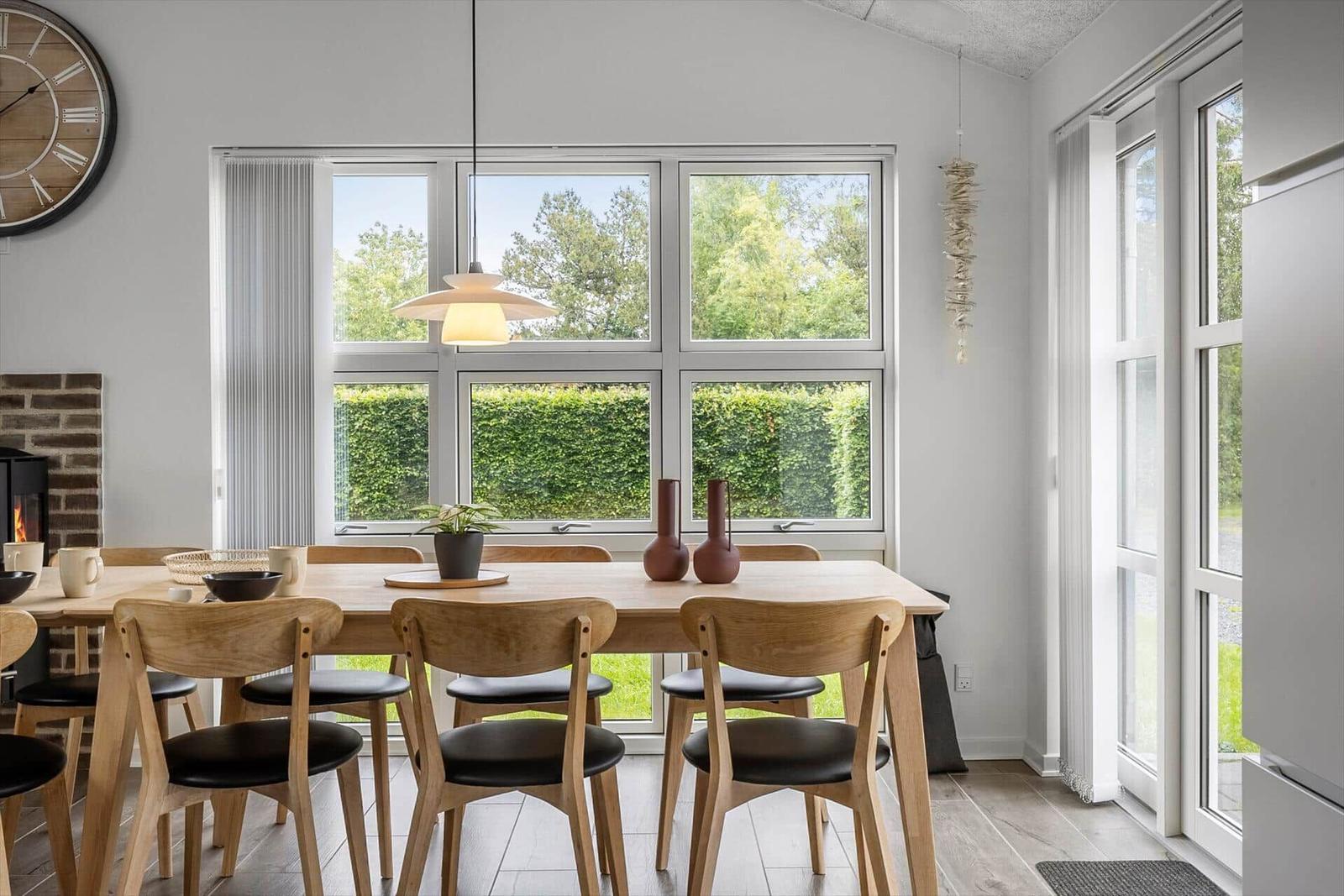 Dining room with wooden table, chairs, and large windows overlooking green outdoors.