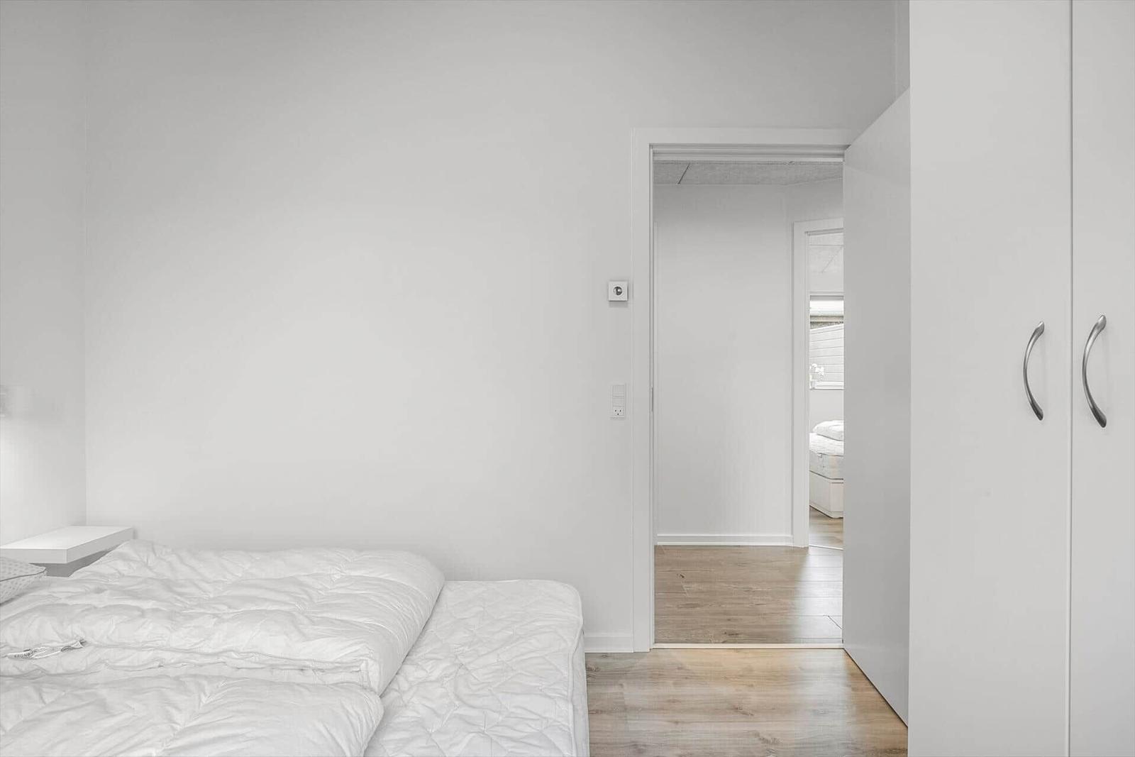 Bedroom with white bed and wardrobe. Passage to another room with wooden floor.
