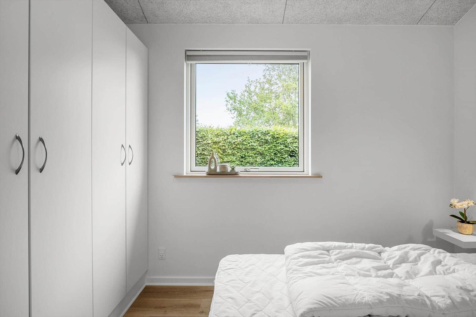 Bedroom with white bed, wardrobe, and window with view of greenery.