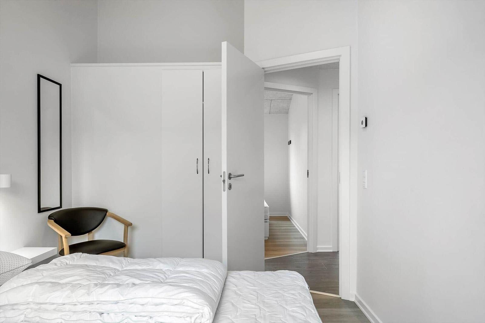 Bedroom with white bed, wardrobe, and chair. Door leads to hallway.