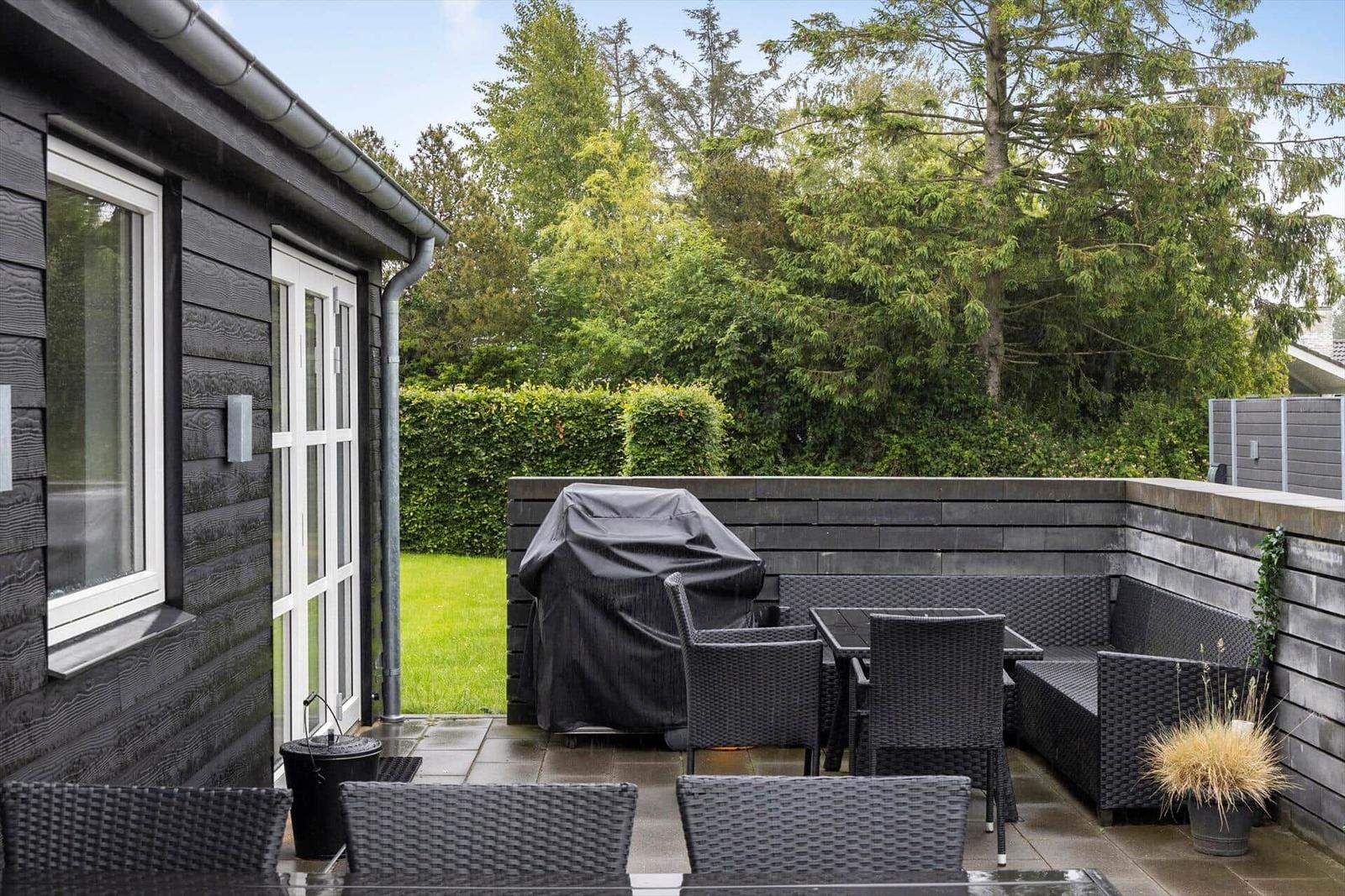 Terrace with seating, grill, and greenery. Black wooden wall and windows.