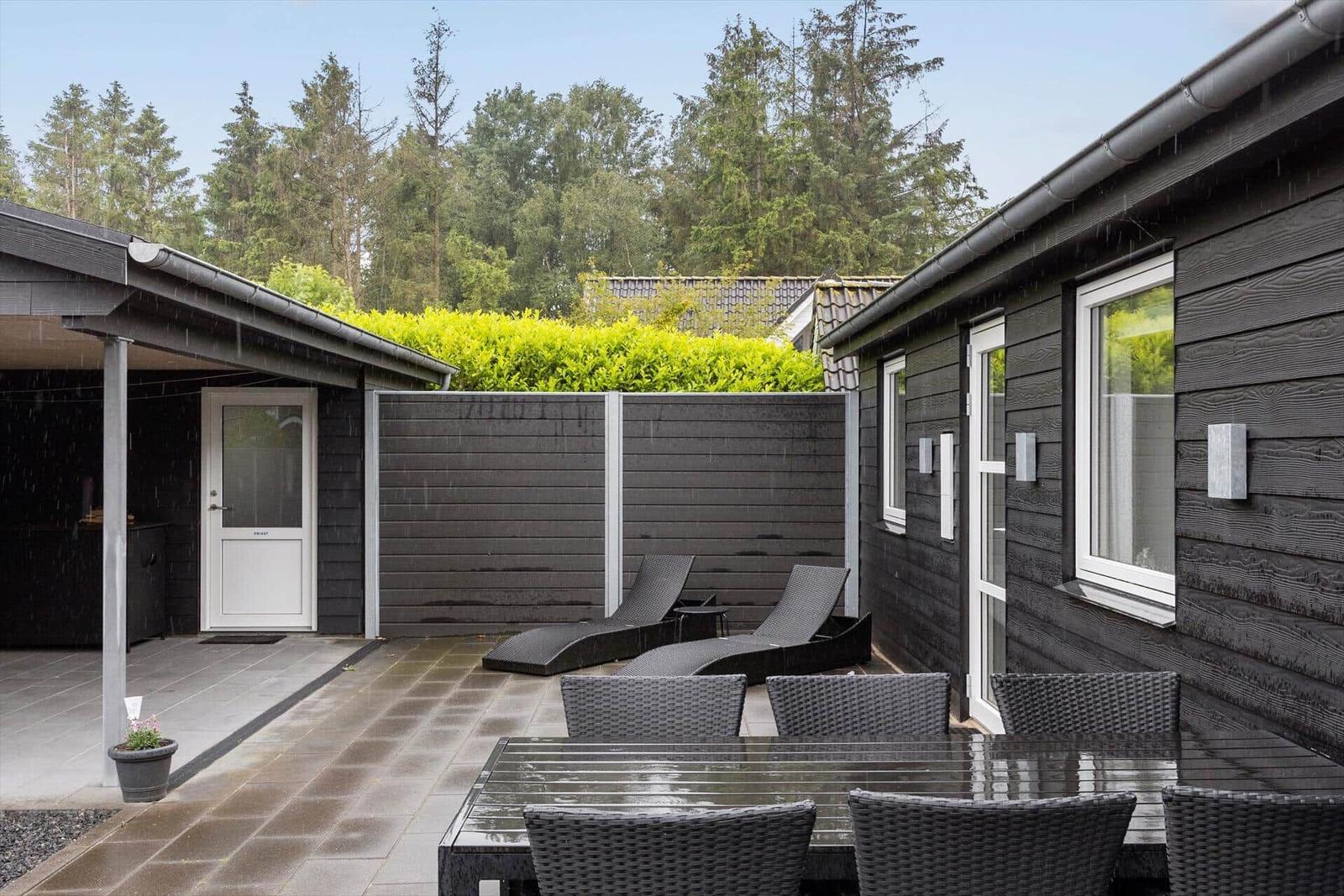 Terrace with table, chairs, and loungers. Dark wood facade and green hedge in the background.