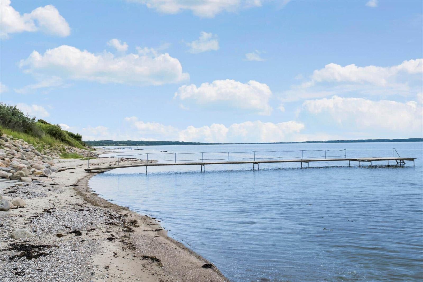 Hafen mit Steg an ruhigem Wasser und Strand mit Steinen und Gras.