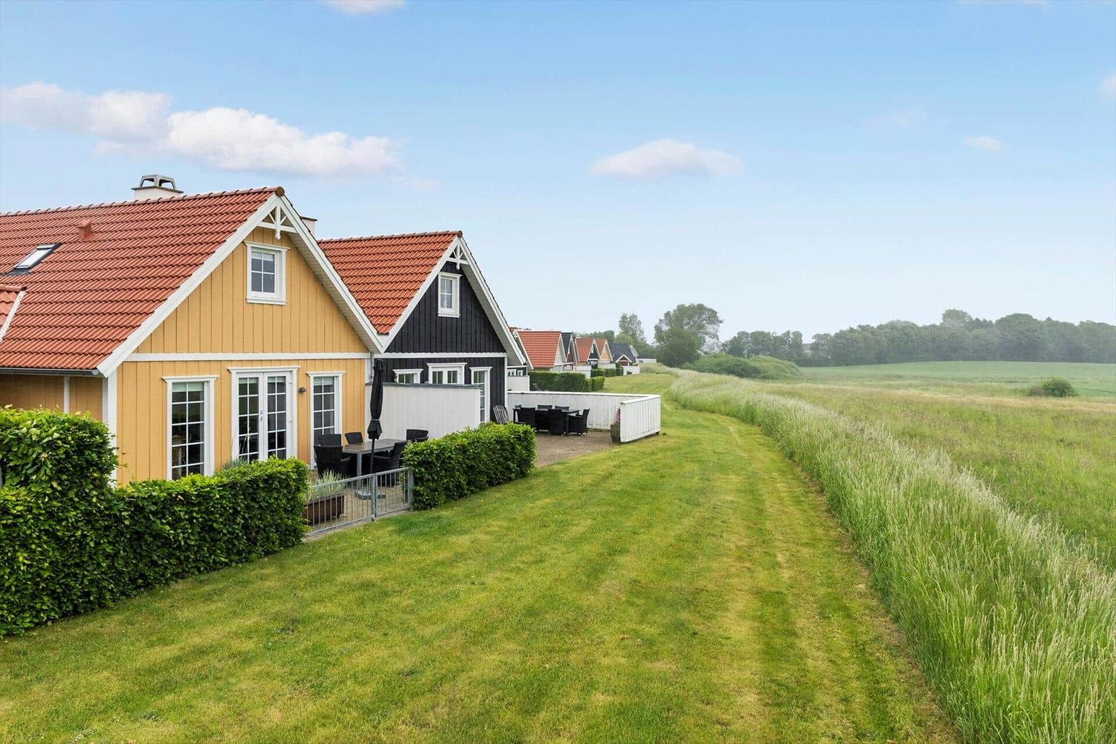 Haus mit Terrasse und Garten, umgeben von Feldern und Bäumen unter blauem Himmel.