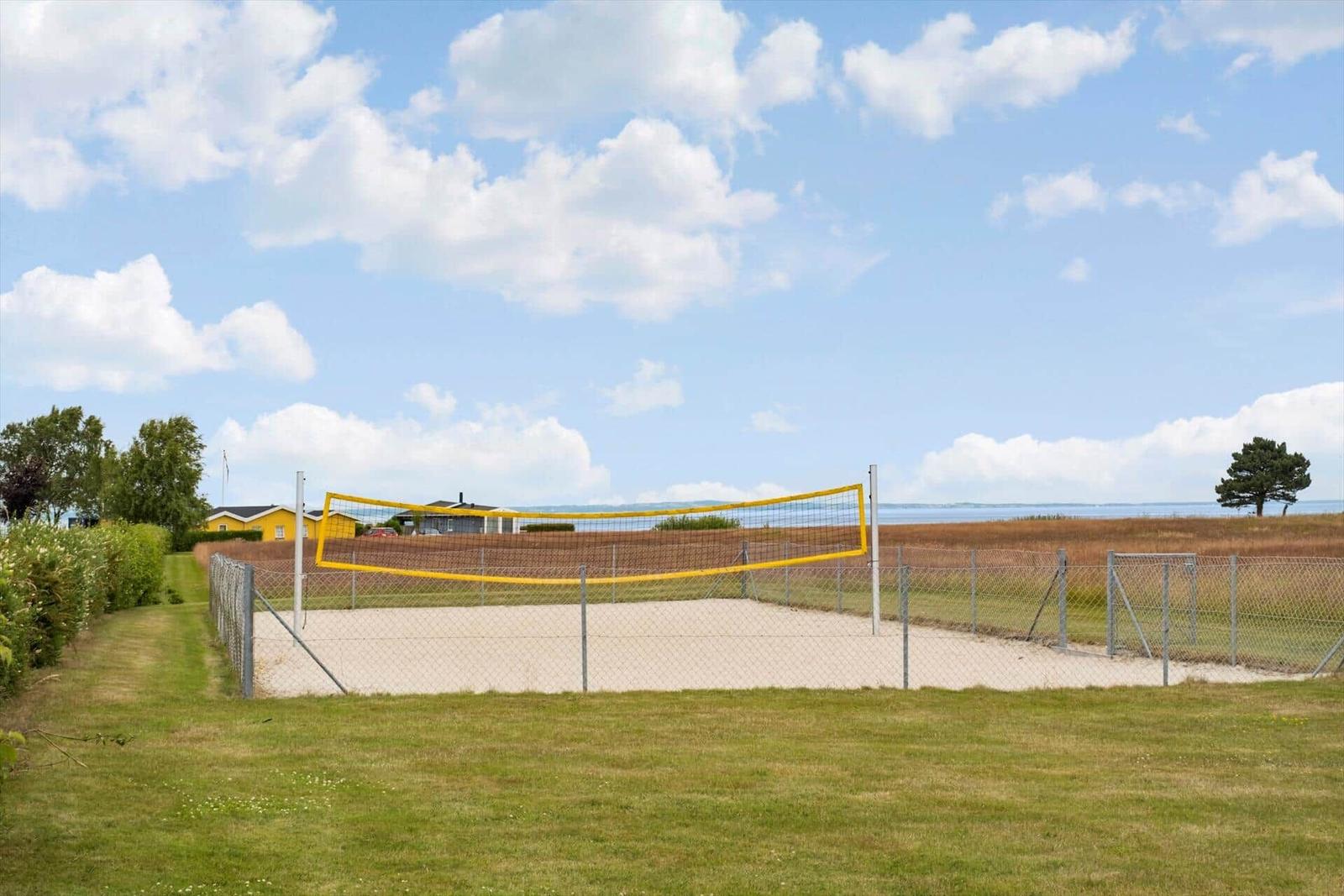 Volleyballplatz mit Sandfläche und gelbem Netz. Umzäunt und grüne Wiesen im Hintergrund.