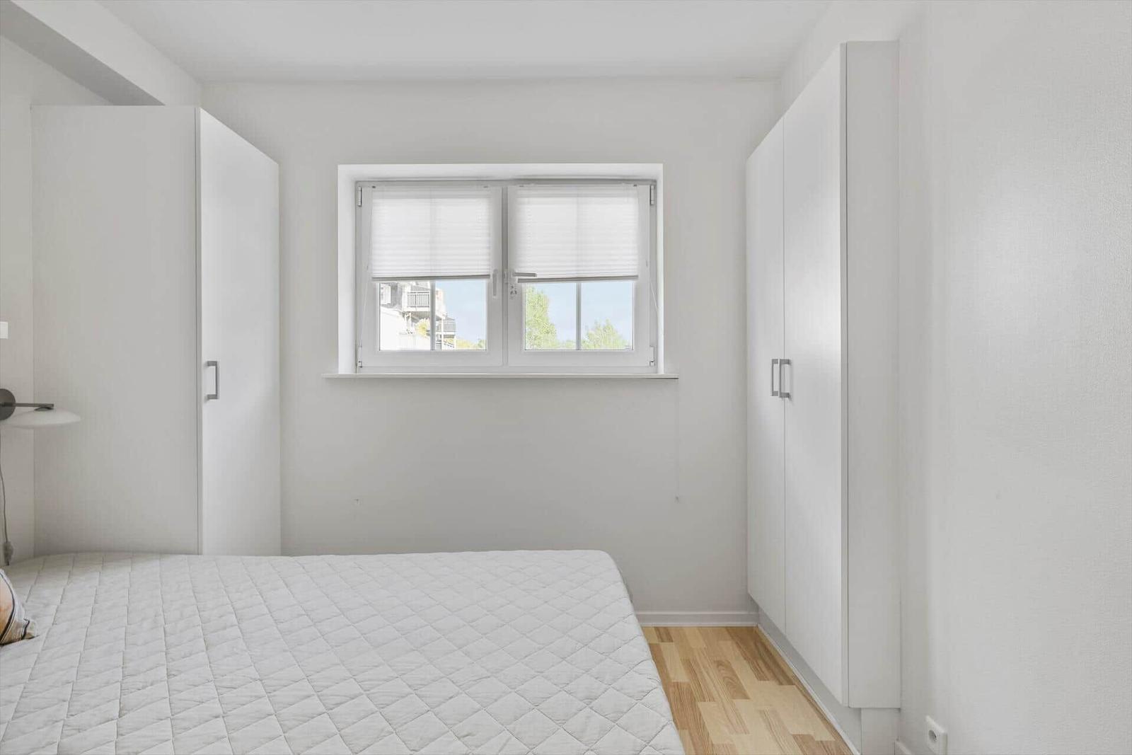 Bedroom with white bed, window, and white wardrobes.