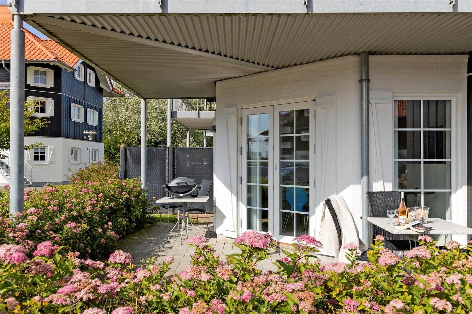 Terrace with grill, table, and chairs, surrounded by pink flowers and a white house.