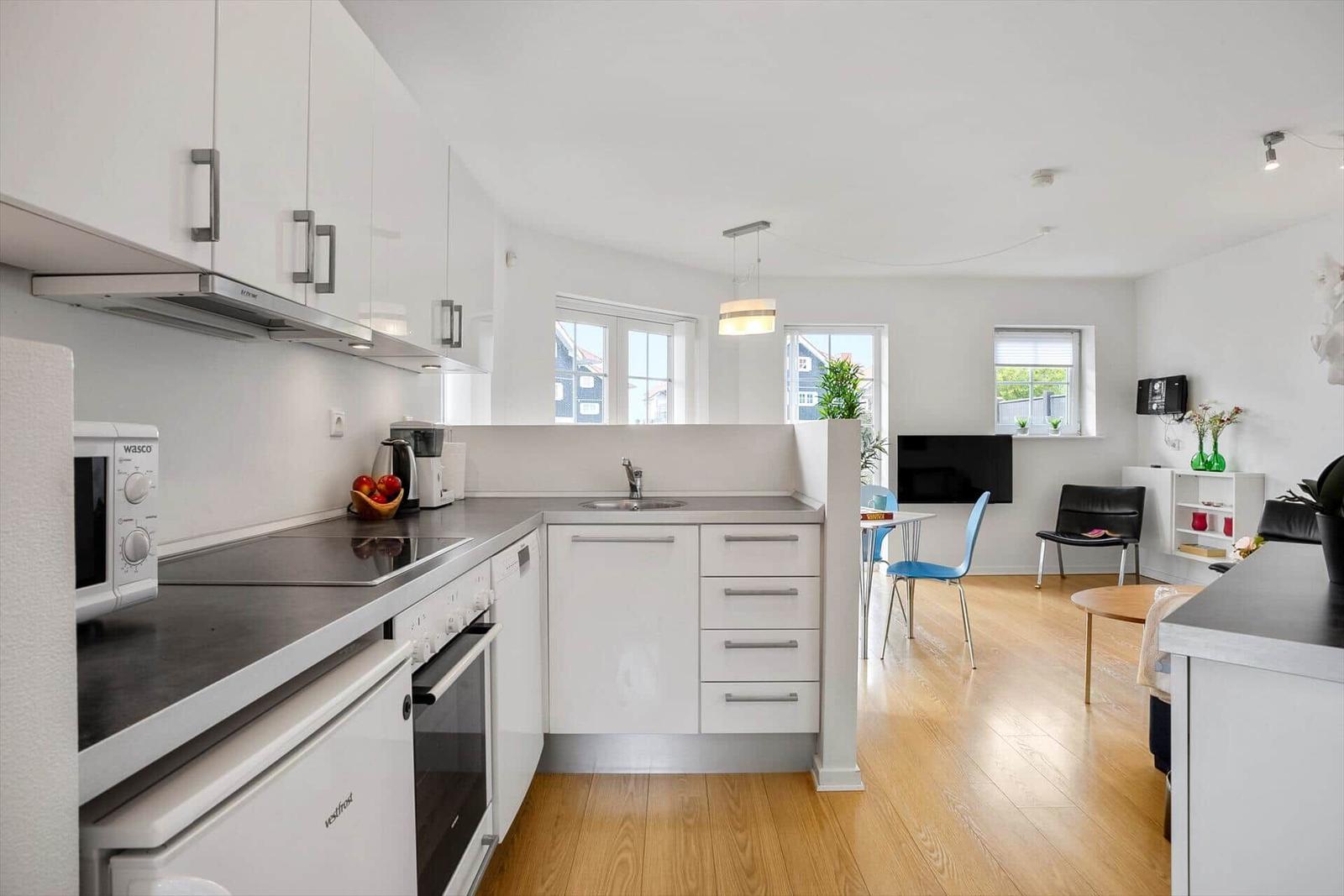 Kitchen with worktop, sink, and appliances. Living area with TV, chairs, and windows.