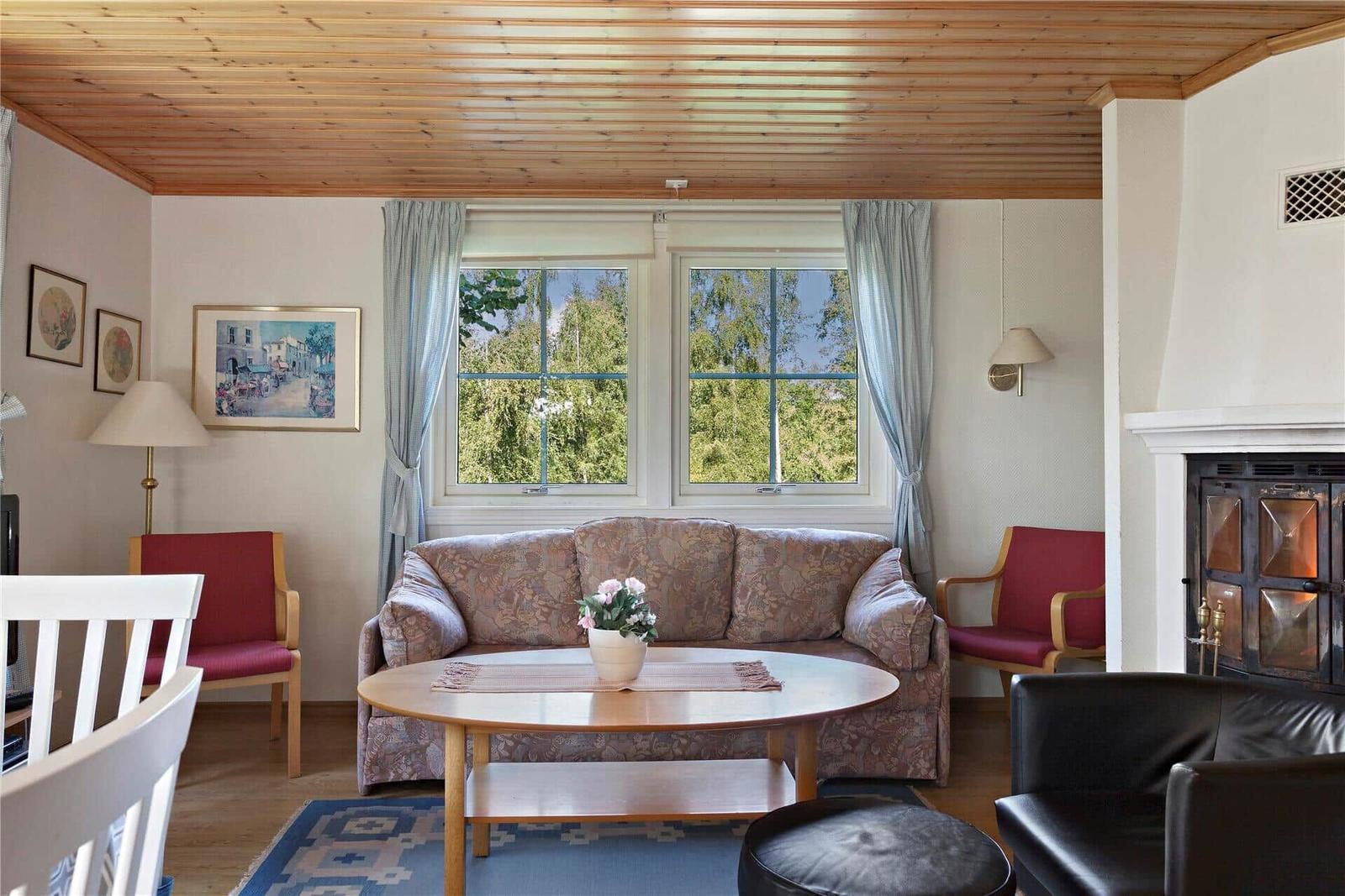 Living room with sofa, table and fireplace. Windows show green trees.
