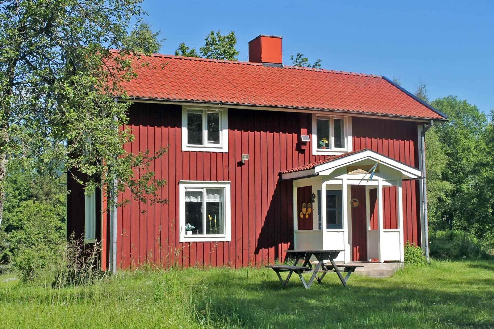 Red wooden house with white windows and porch. Surrounded by greenery with picnic table.