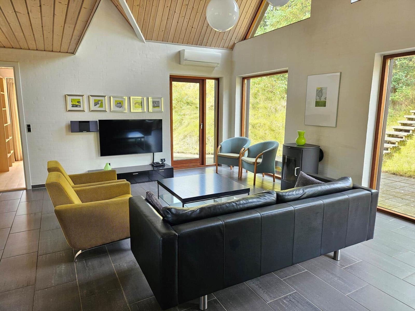 Wohnzimmer mit Sofa, Sesseln und Fernseher. Große Fenster und Holzdecke.