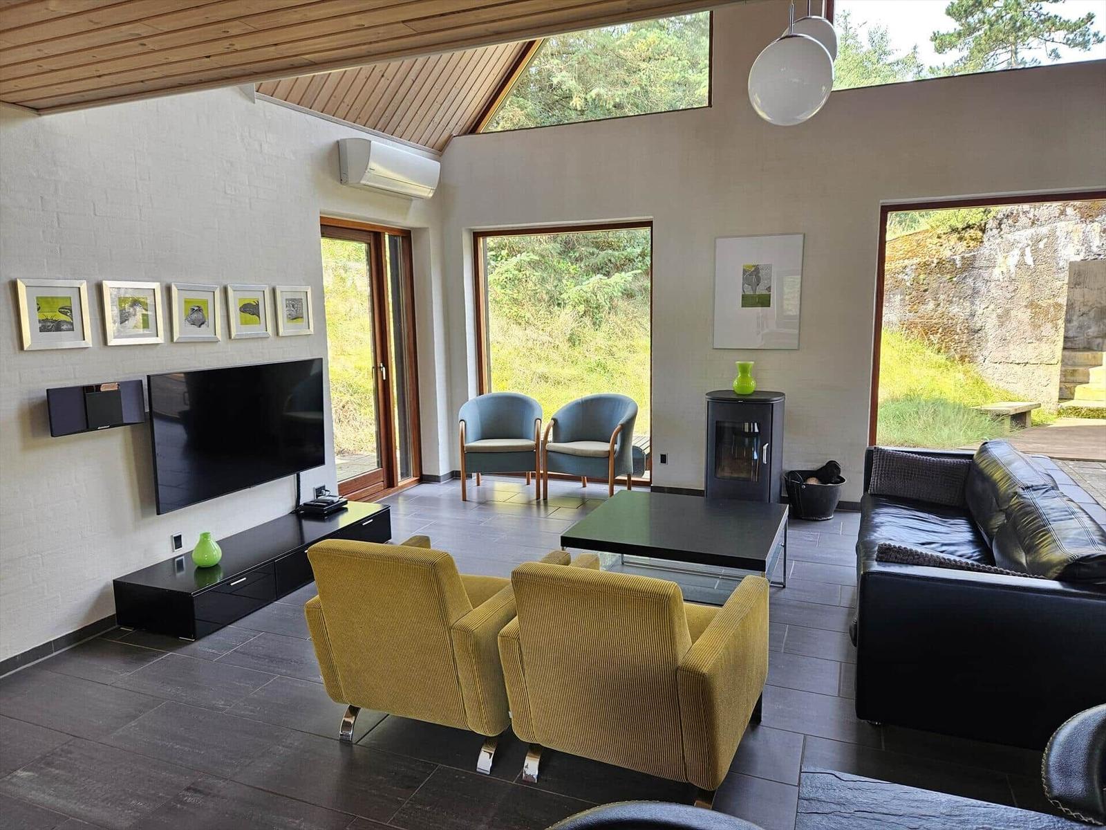 Wohnzimmer mit Fernseher, Sofa und Sesseln. Große Fenster und Holzdecke.