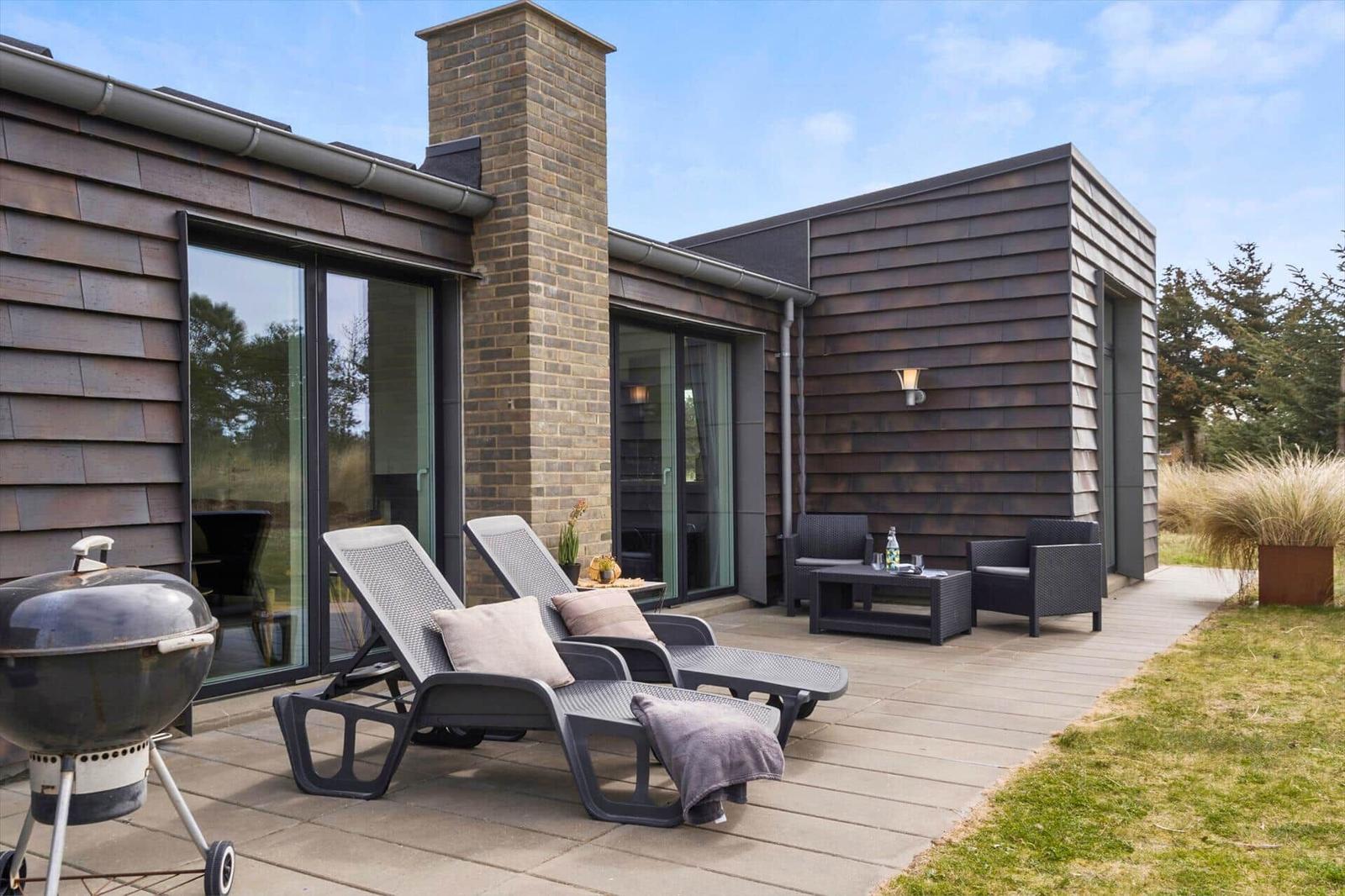 Terrasse mit Liegen, Sitzgruppe und Grill. Modernes Haus mit Holzfassade und Glasfront.