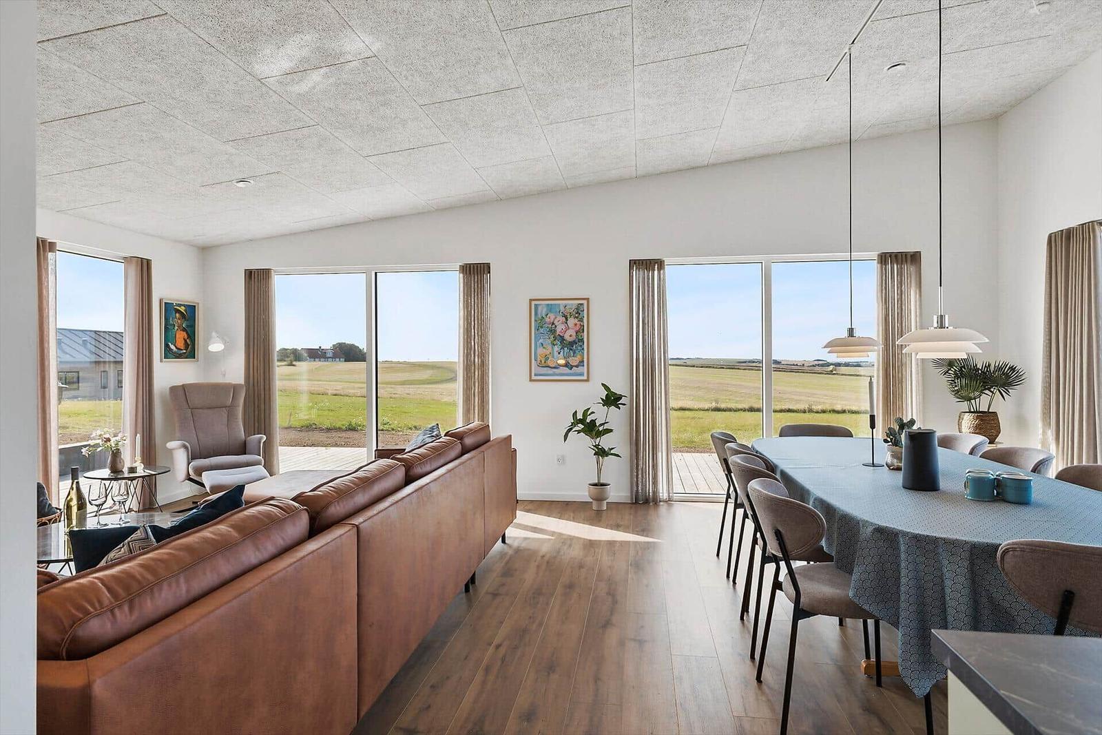 Wohn- und Essbereich mit braunem Sofa, Tisch und großen Fenstern zur Landschaft.